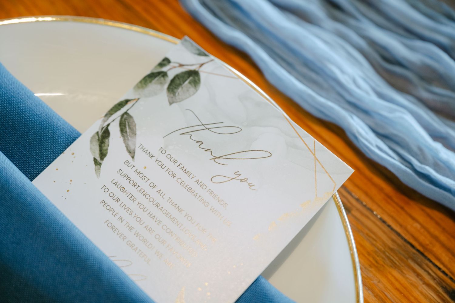 Wedding invitation suite with watercolor greenery details laid out on a wooden table with blue fabric accents. - The Brix on the Fox