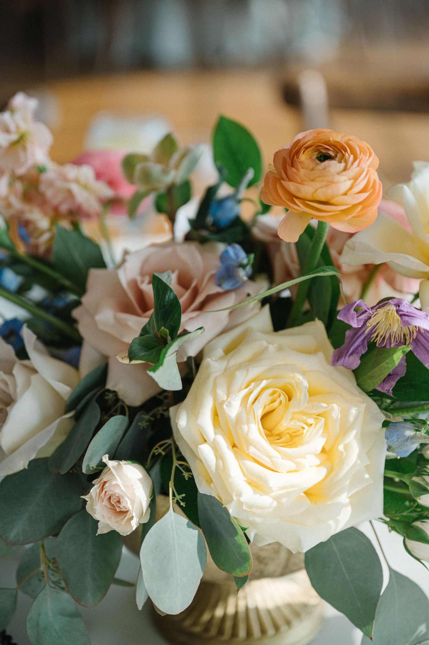 A romantic floral arrangement featuring cream roses, peach ranunculus, and eucalyptus leaves in a gold vase.