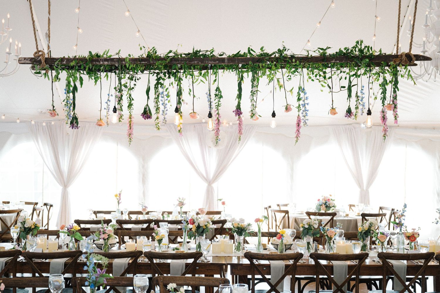 Elegant wedding reception tent with hanging greenery installation and soft pink floral accents above dining tables.
