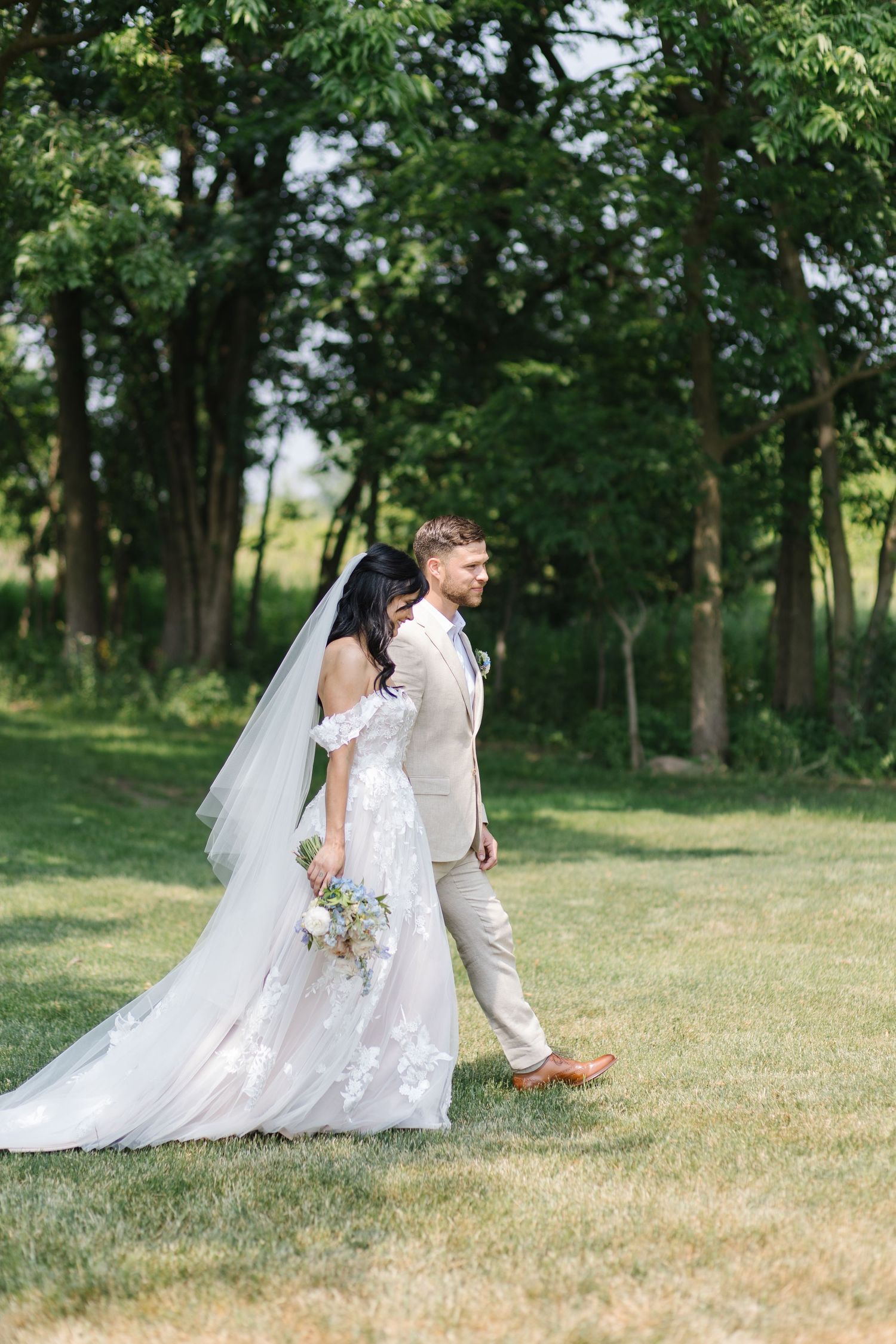 A couple walks together on grassy lawn with trees in background on their wedding day, wearing white attire.