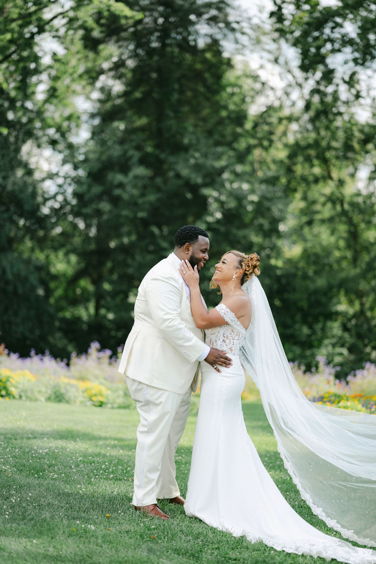 A romantic wedding photo of newlyweds in all white embracing on a manicured lawn with lush greenery. - Bird Haven Greenhouse