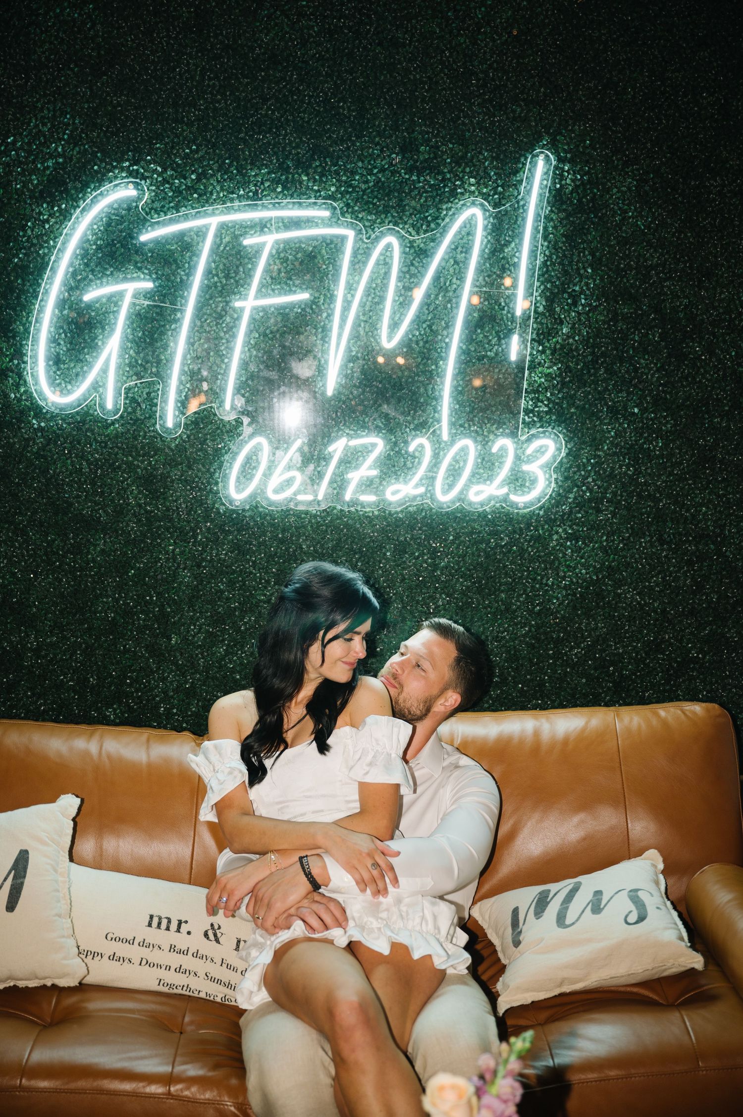 Neon GTFM sign glows above a brown leather couch with decorative Mr & Mrs pillows in a romantic setting.