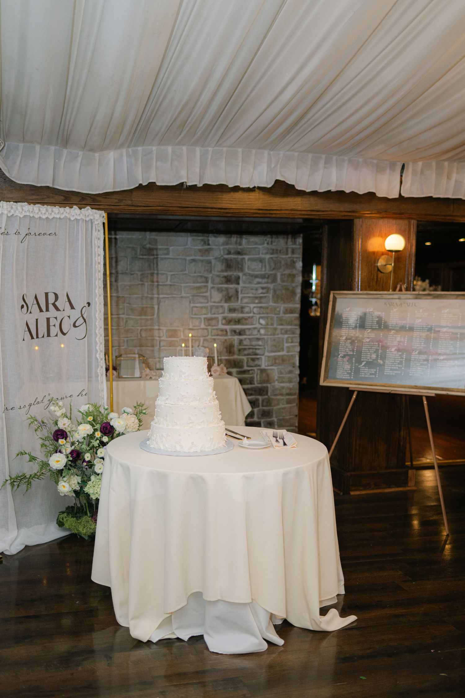 Three elegant white wedding cakes displayed on round tables with white linens in a rustic brick-walled reception venue. - Meyer's Castle