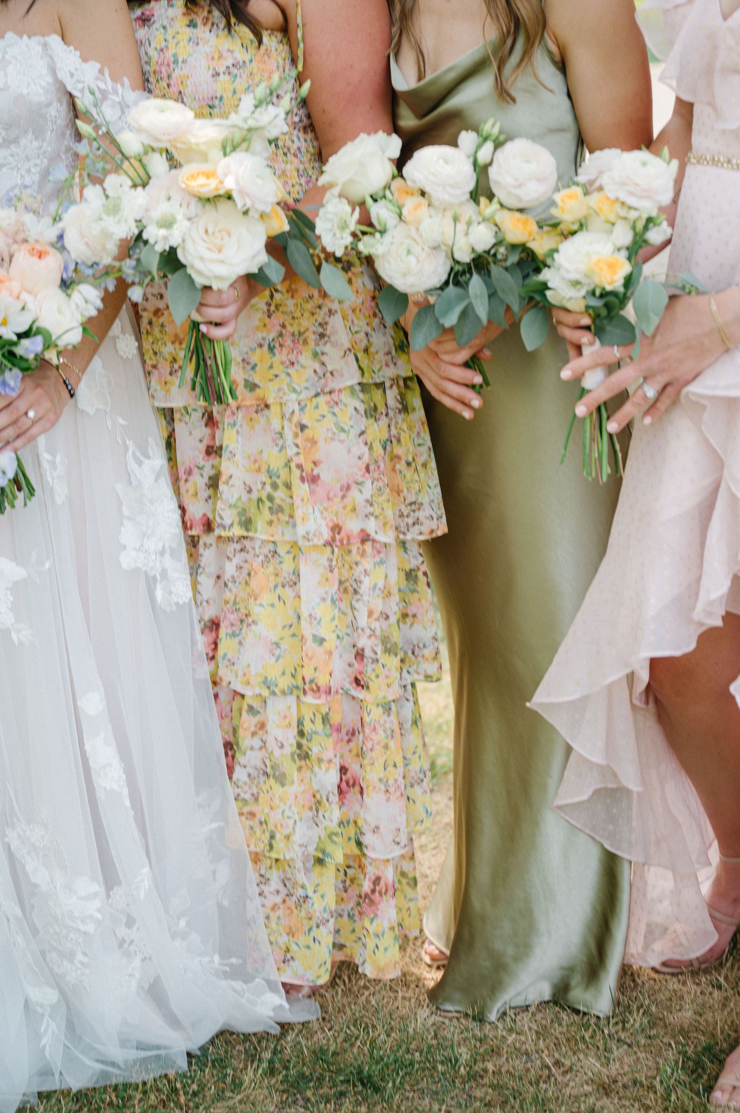 Wedding party members hold white floral bouquets while wearing dresses in sage green, floral print, and white styles.