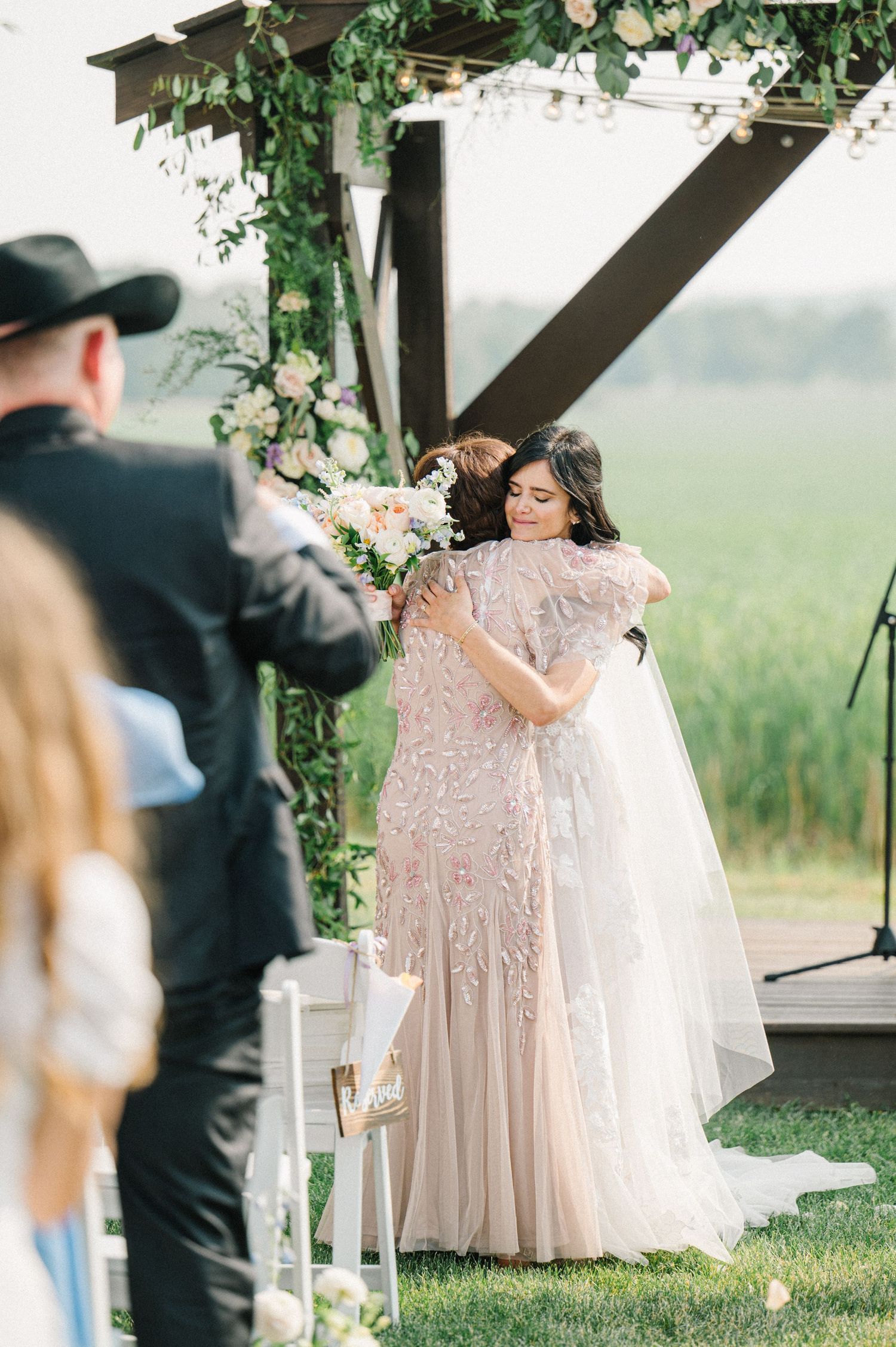 A beautiful outdoor wedding ceremony takes place under a white arbor decorated with greenery and florals in a rustic setting.