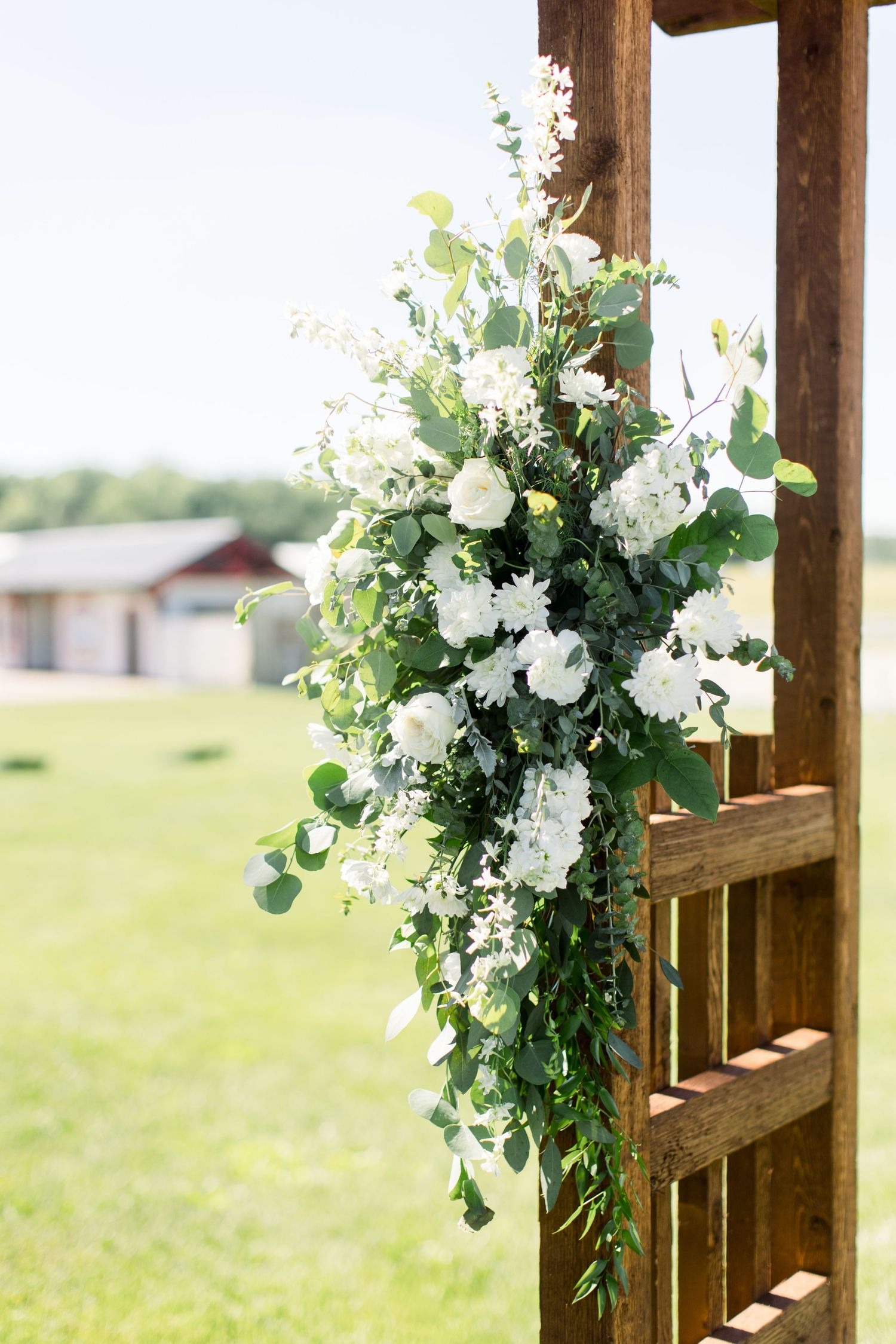 Elegant white floral arrangement with greenery mounted on wooden post at outdoor wedding venue.