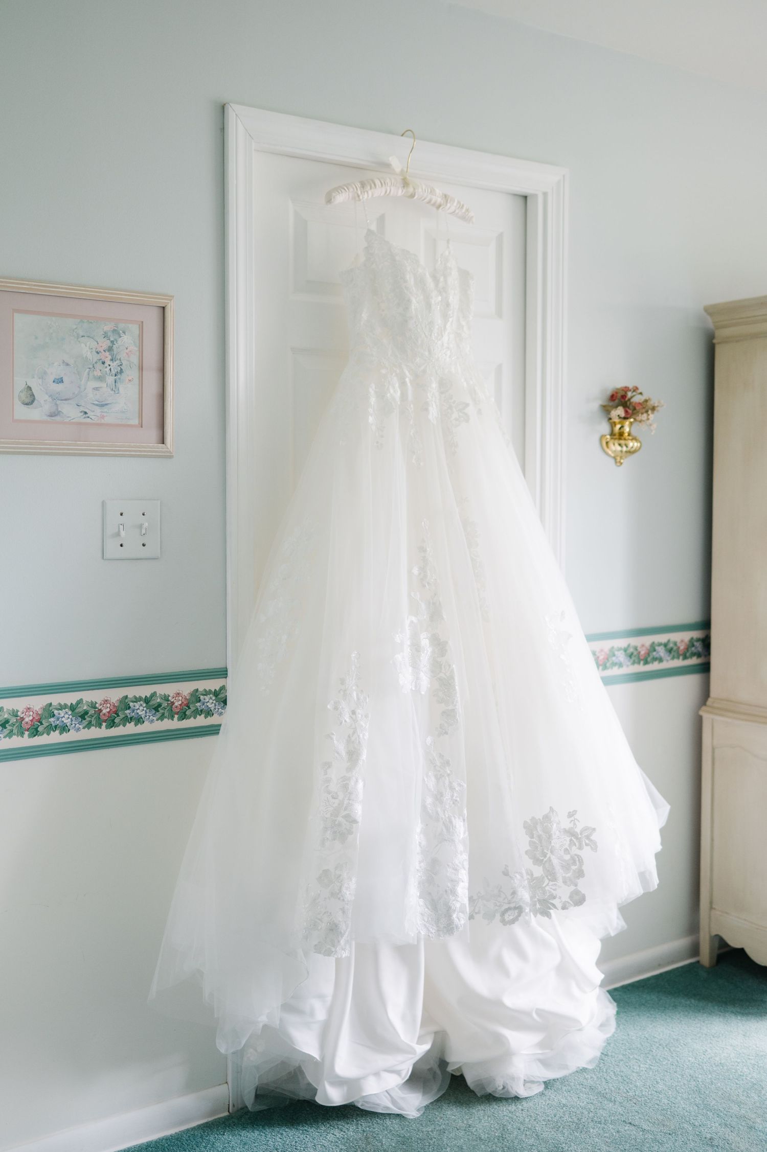 A white lace wedding gown with intricate details hangs elegantly against a light blue wall.
