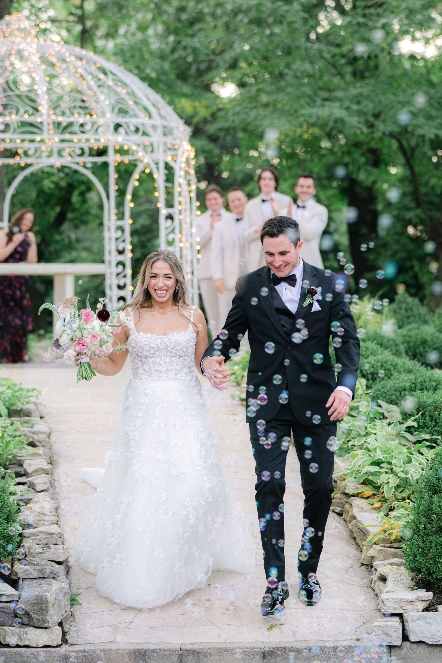 Couple walks along garden path surrounded by floating bubbles at outdoor wedding venue with white gazebo and greenery. - Meyer's Castle