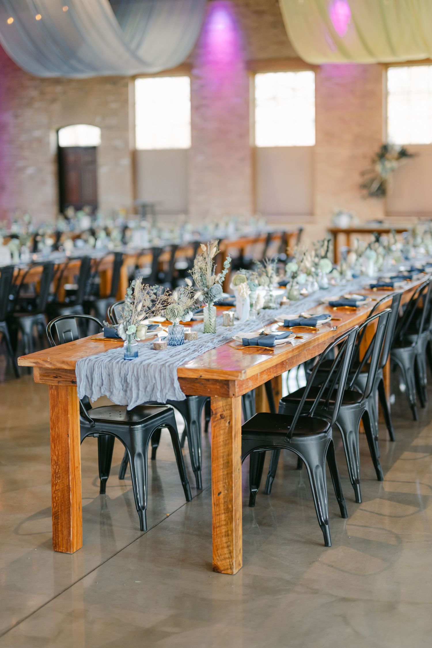 Long farm tables with black metal chairs and gray table runners set up for an elegant industrial wedding reception. - The Brix on the Fox
