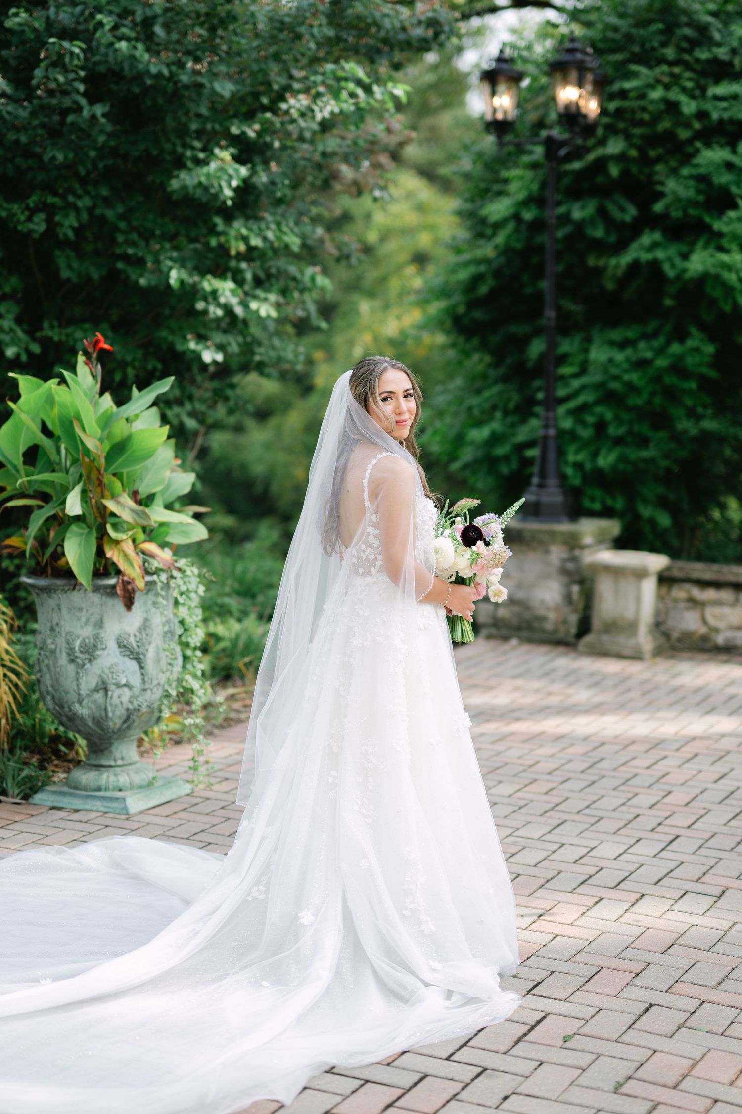 A bride in a flowing white wedding gown and veil poses gracefully on a brick paved garden path. - Meyer's Castle Events
