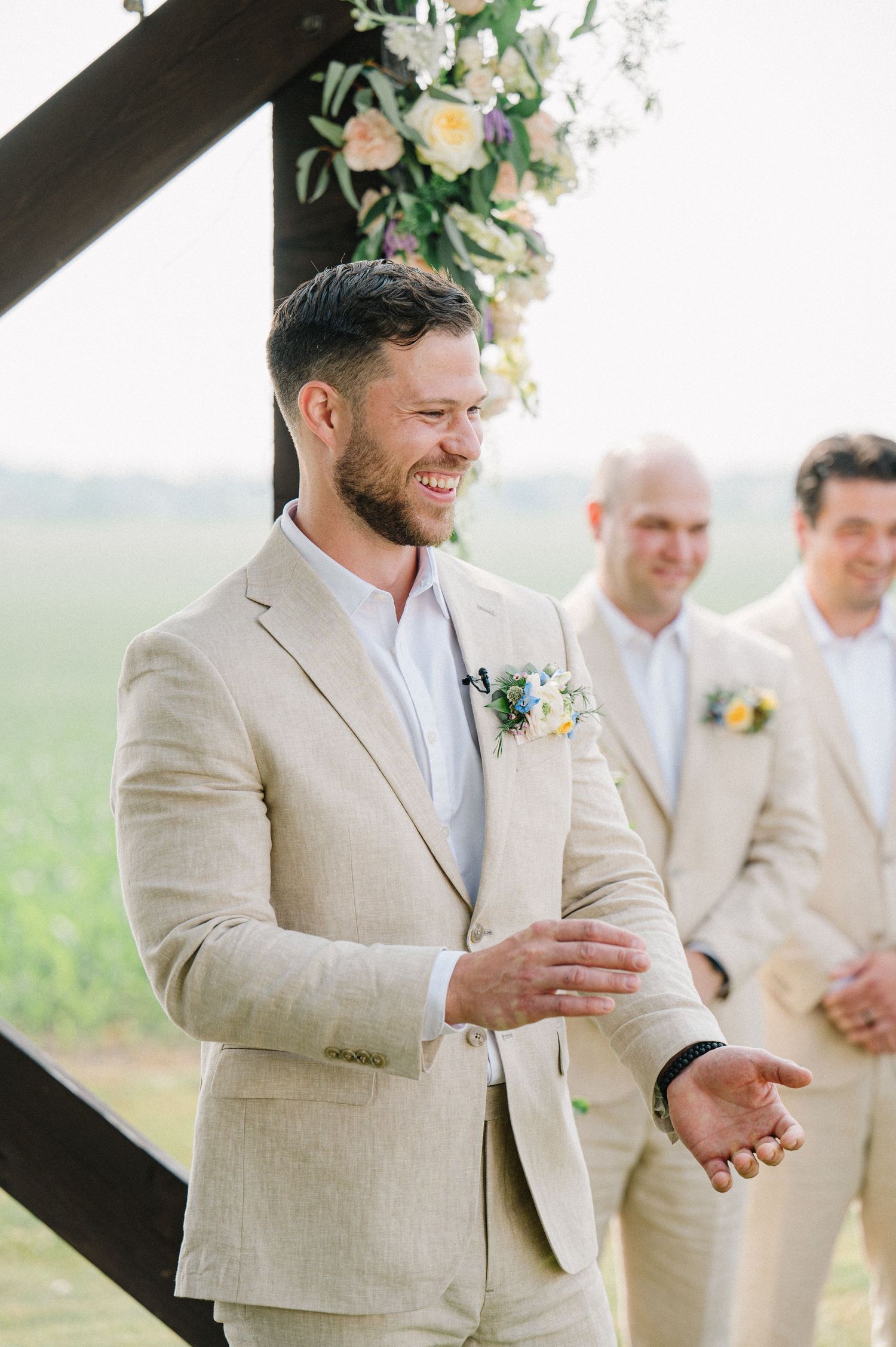 A groom laughs joyfully at an outdoor wedding ceremony wearing a light beige suit and boutonniere.