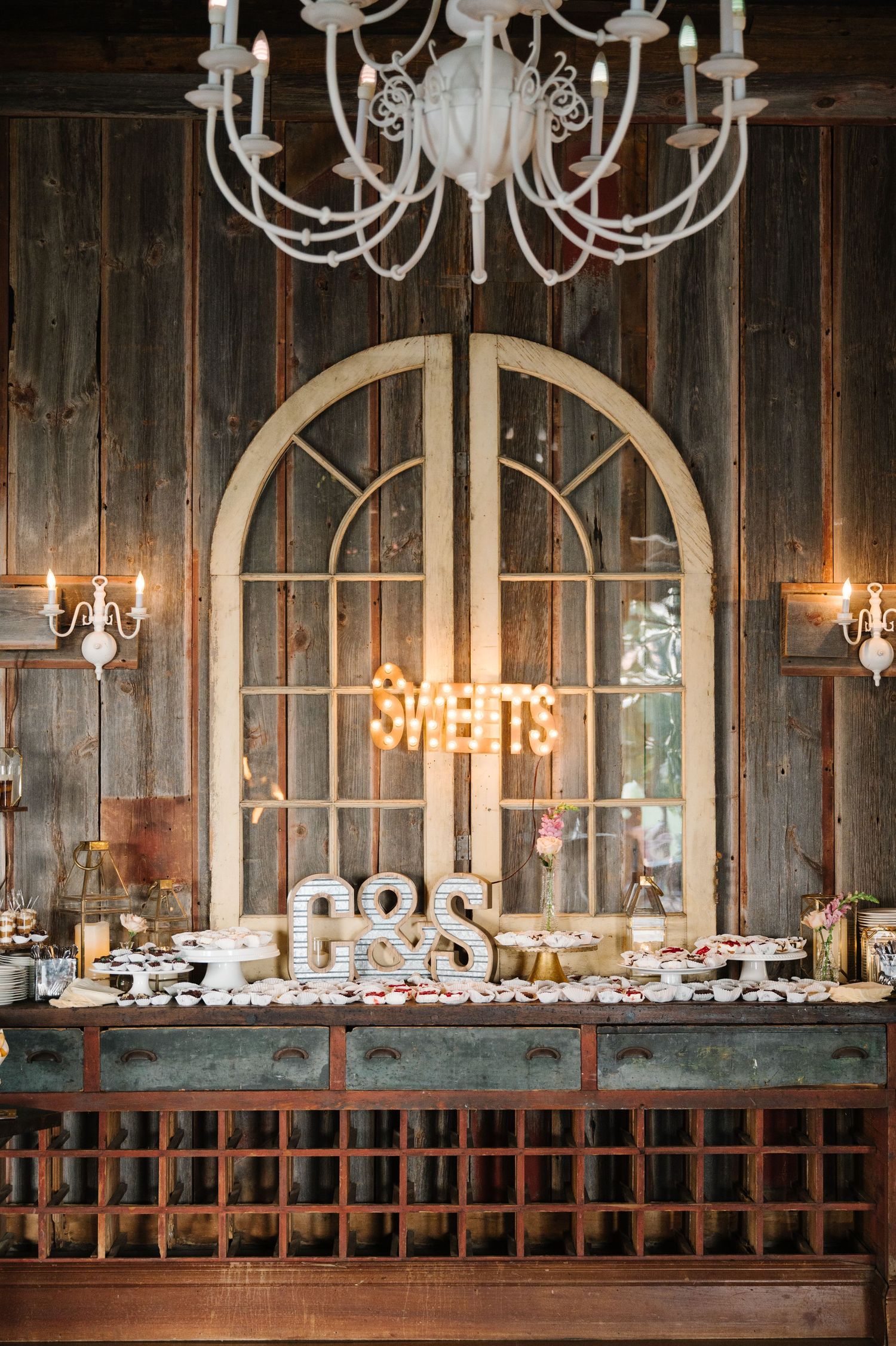Rustic dessert table with illuminated SWEETS sign and white chandelier above vintage wooden arched doors and wall.