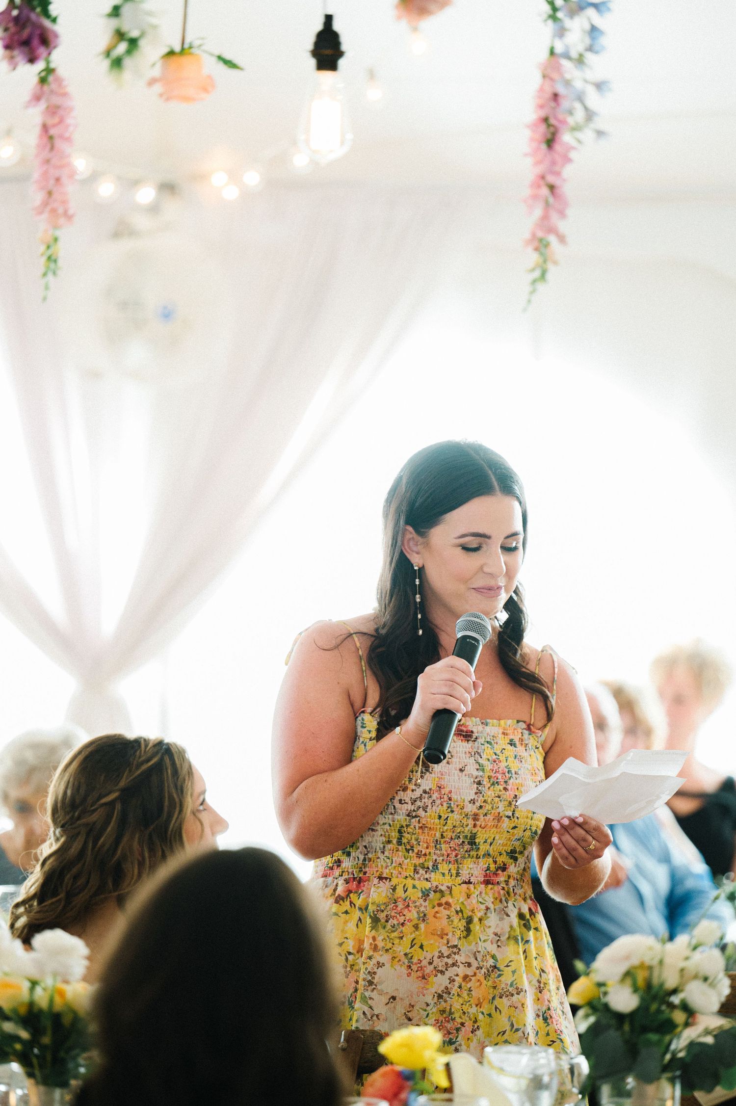 A bridesmaid in a floral yellow dress delivers a heartfelt speech at an outdoor wedding reception under floral decorations.