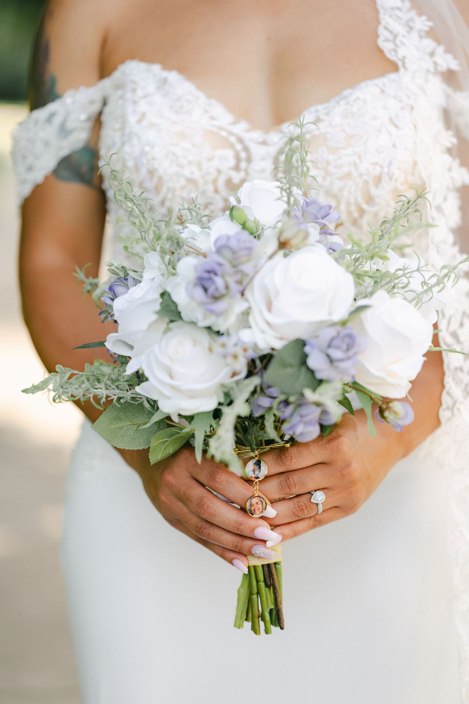 A delicate bridal bouquet with white roses and pale purple flowers held by hands wearing sparkly engagement rings. - Bird Haven Greenhouse