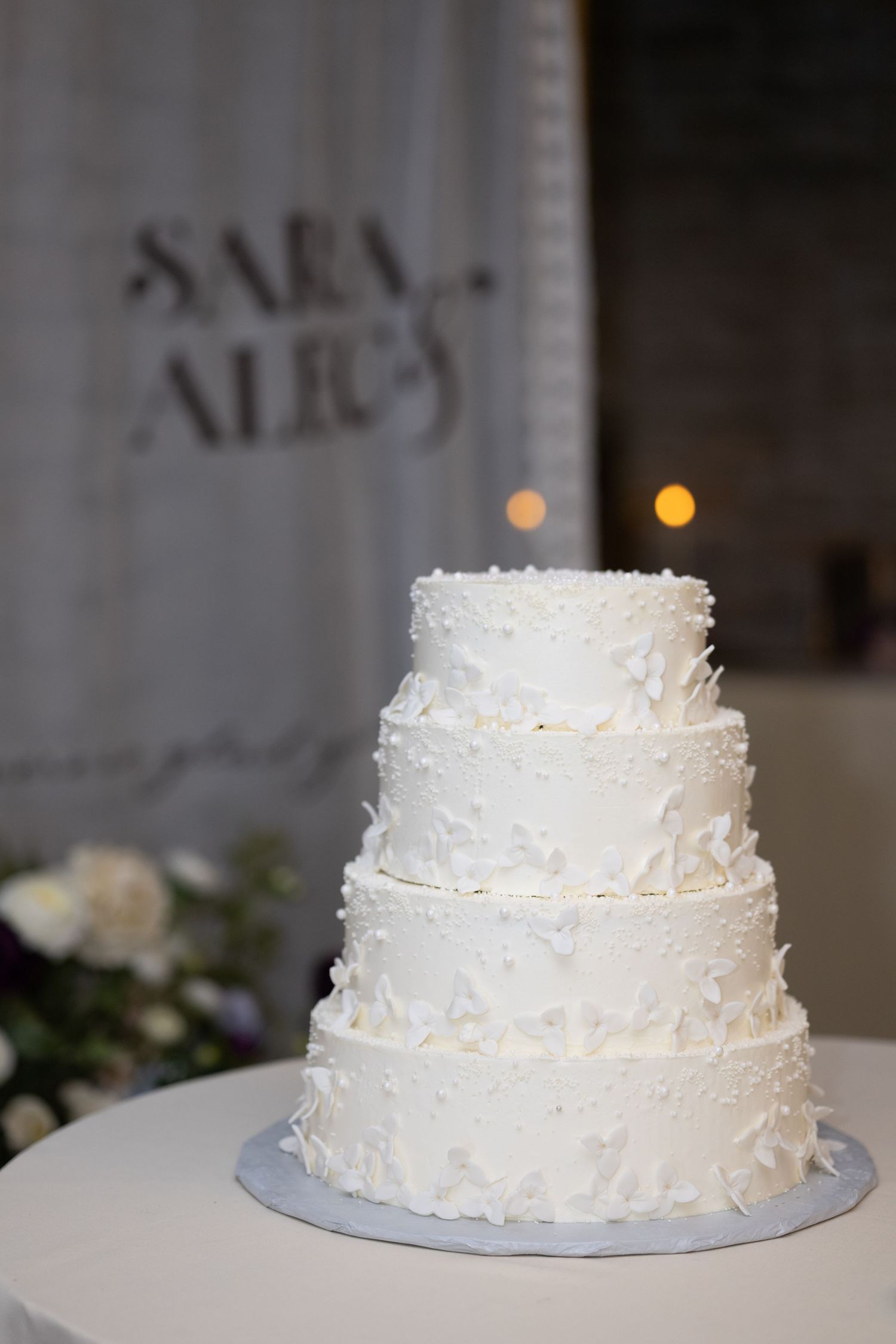 A four-tier white wedding cake with textured frosting and glowing lights in the background.
