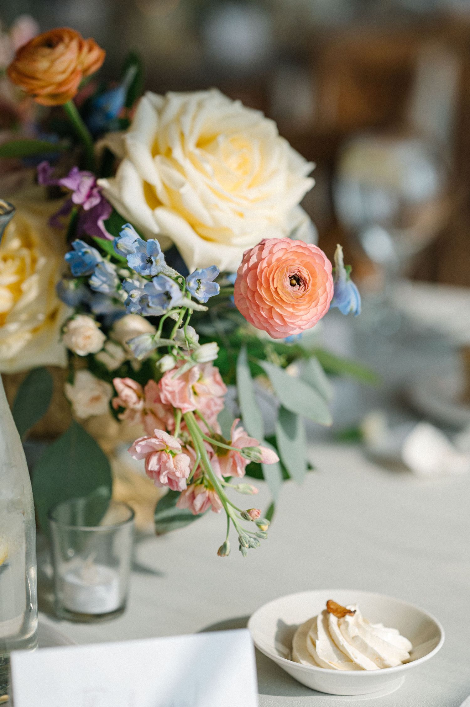 Delicate floral arrangement with cream roses, coral ranunculus, and blue forget-me-nots next to whipped dessert on white table.