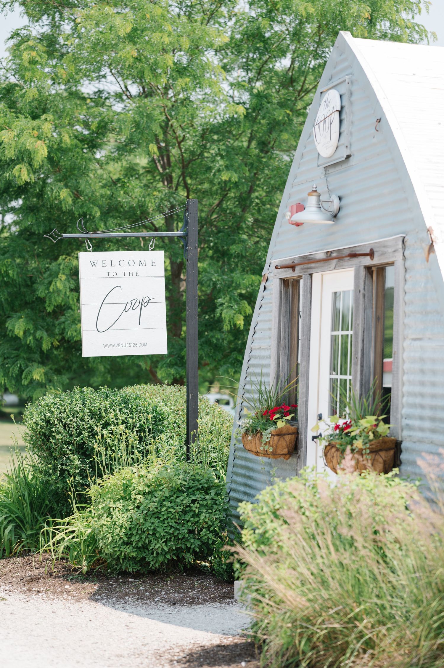 A quaint white metal A-frame building with flower boxes and a welcome sign surrounded by lush greenery.