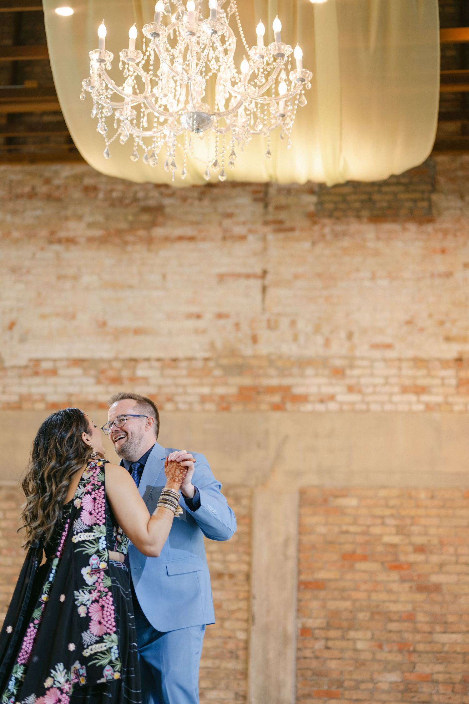 A couple shares a romantic dance moment under elegant crystal chandeliers in a rustic brick-walled venue. - The Brix on the Fox
