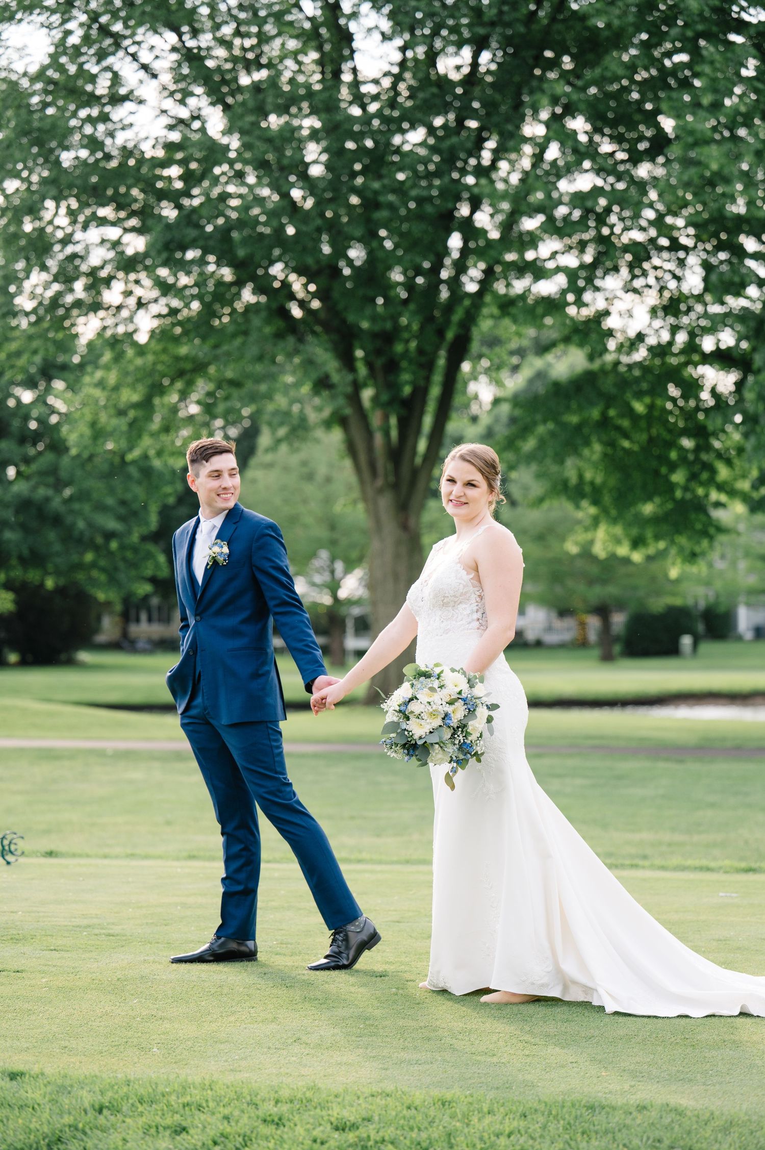 A couple in wedding attire walks hand in hand across a lush green lawn surrounded by tall trees on their special day.