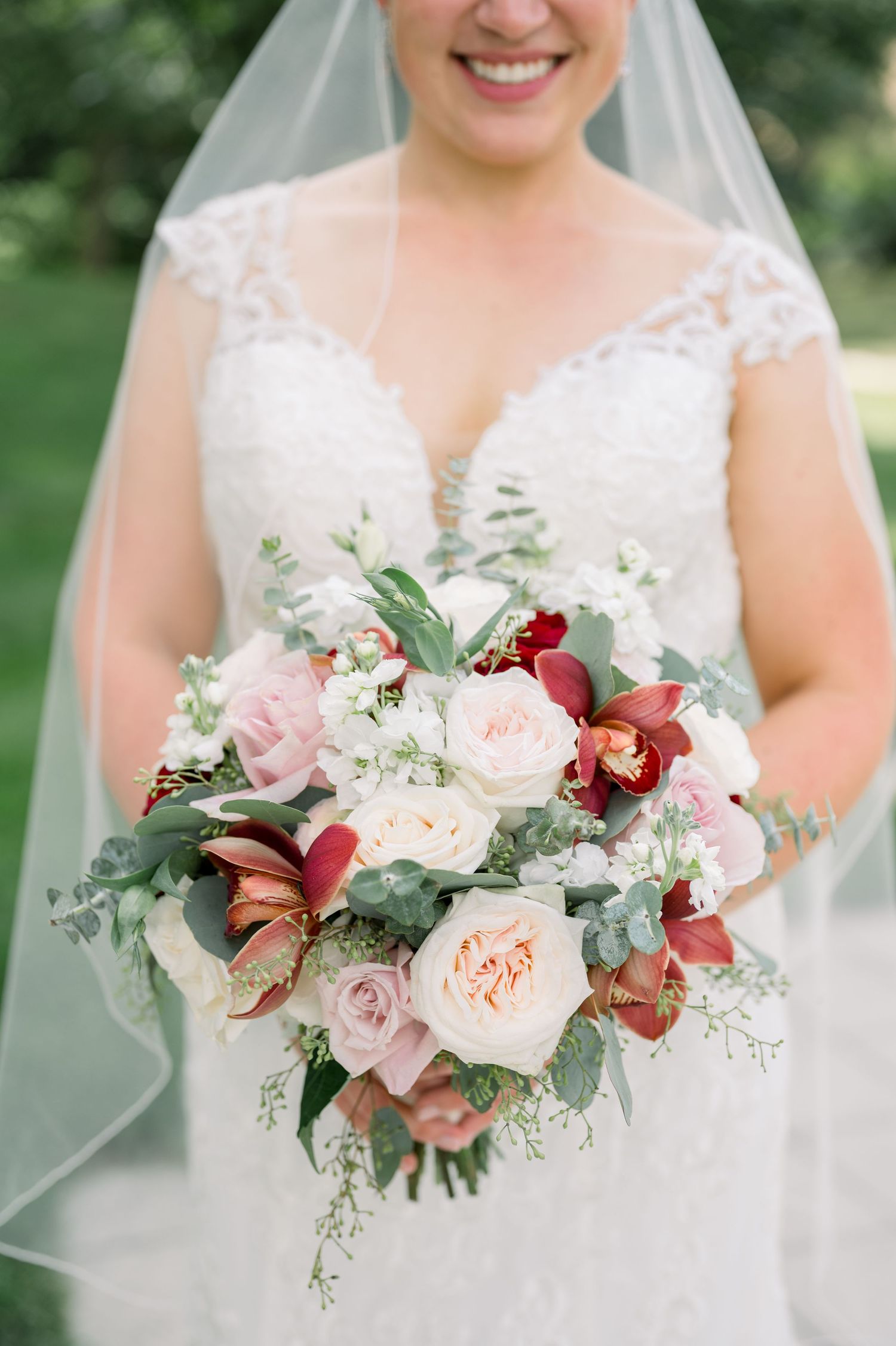 A bridal bouquet features pink garden roses, burgundy orchids and delicate greenery against a white lace wedding dress. - Q Center