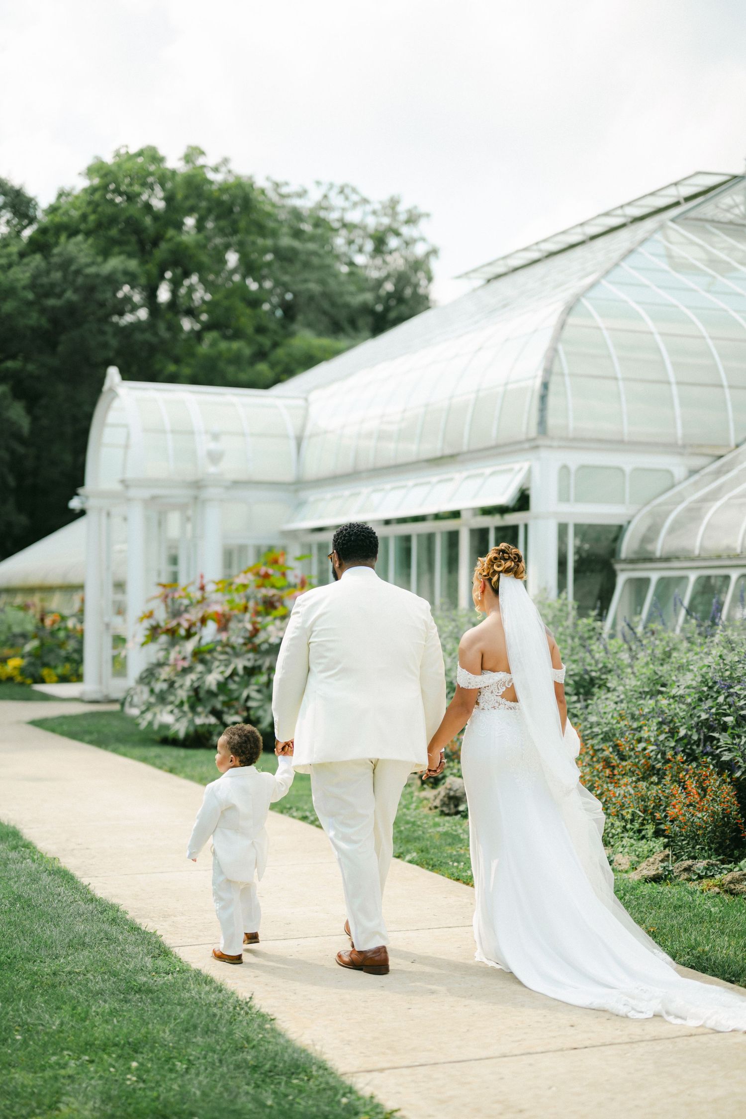 A wedding couple and small child walk along a garden path beside a grand glass conservatory on a sunny day. - Bird Haven Greenhouse