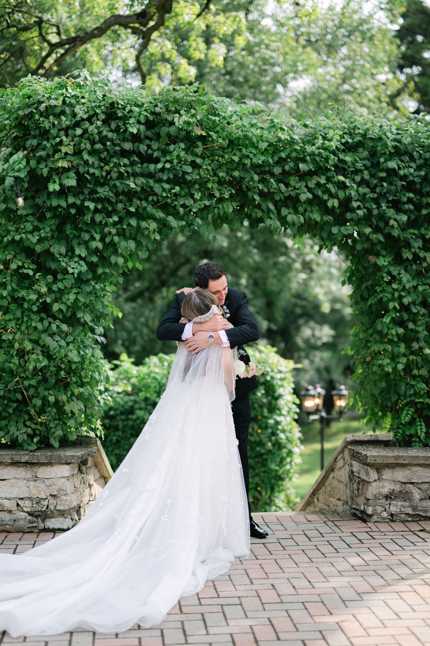 A romantic wedding couple shares a tender embrace on a brick patio surrounded by lush green ivy walls. - Meyer's Castle Events
