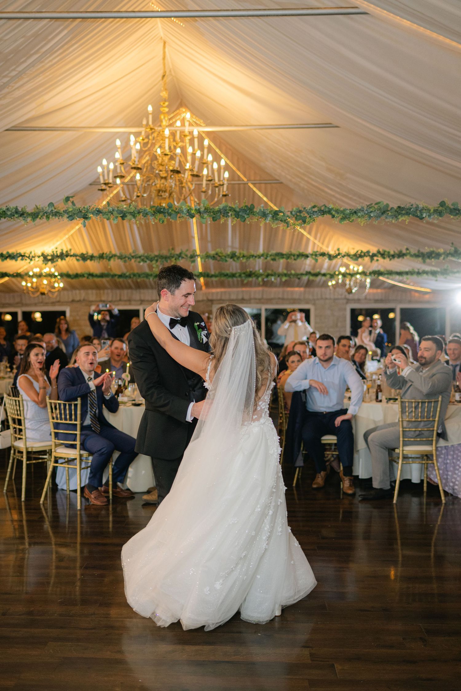 A couple shares their first dance at an elegant wedding reception under a draped tent with a glowing chandelier. - Meyer's Castle Events