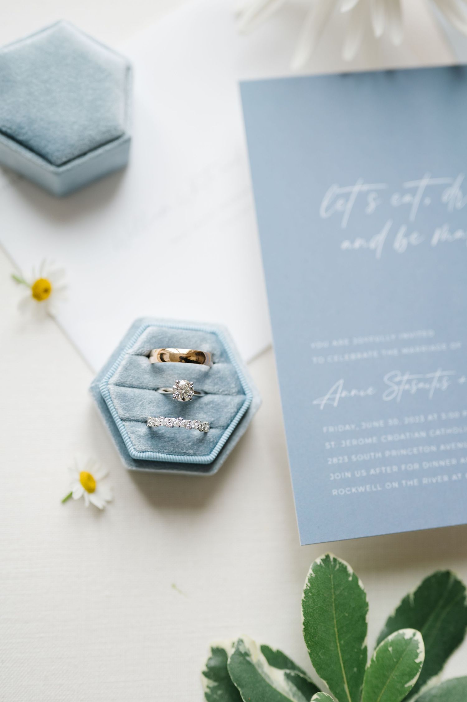 Wedding rings displayed in light blue velvet hexagonal ring box with invitation and daisies.