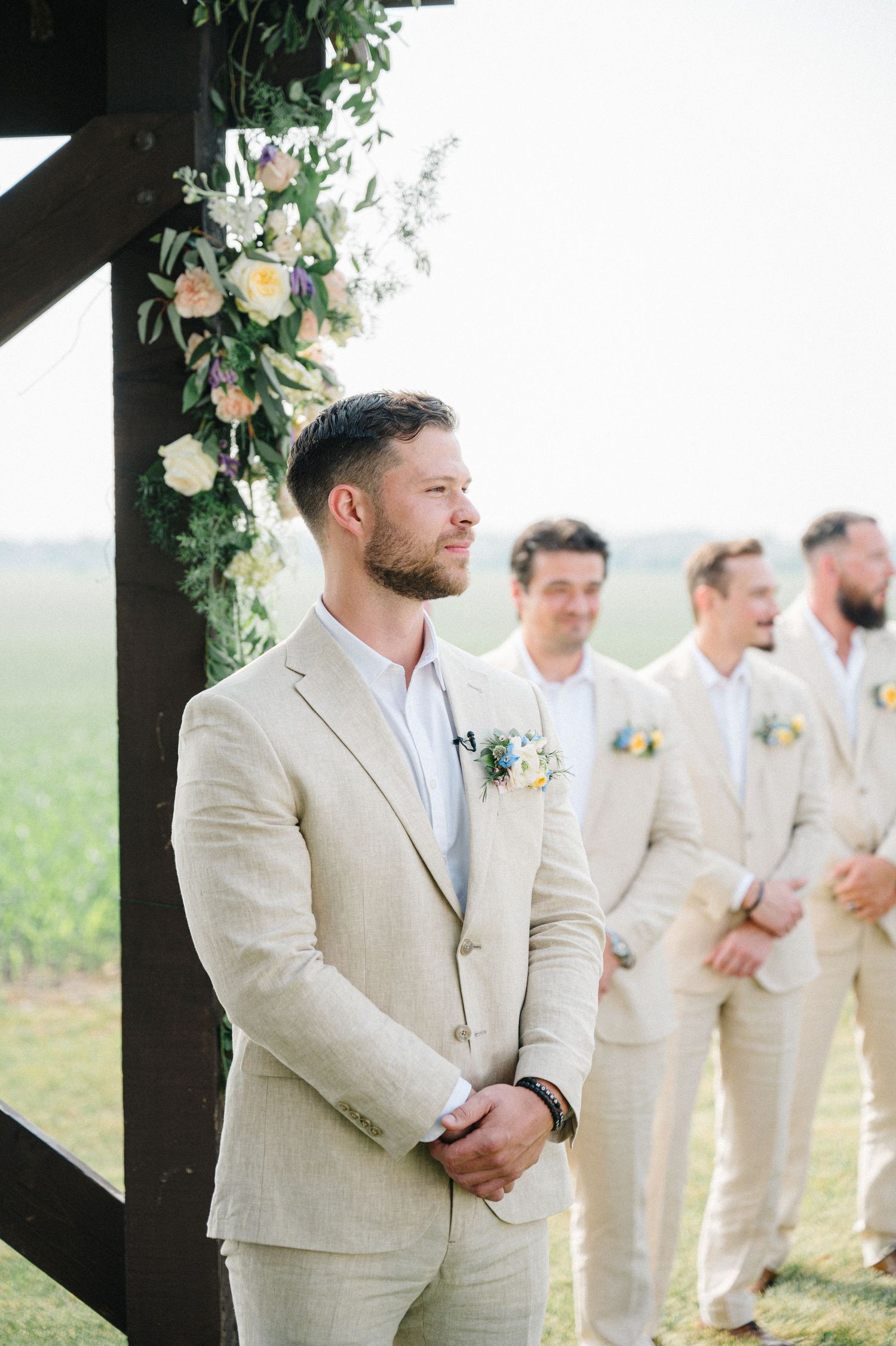 A groomsman in a beige linen suit stands at an outdoor wedding ceremony with floral decorations and other groomsmen behind him.