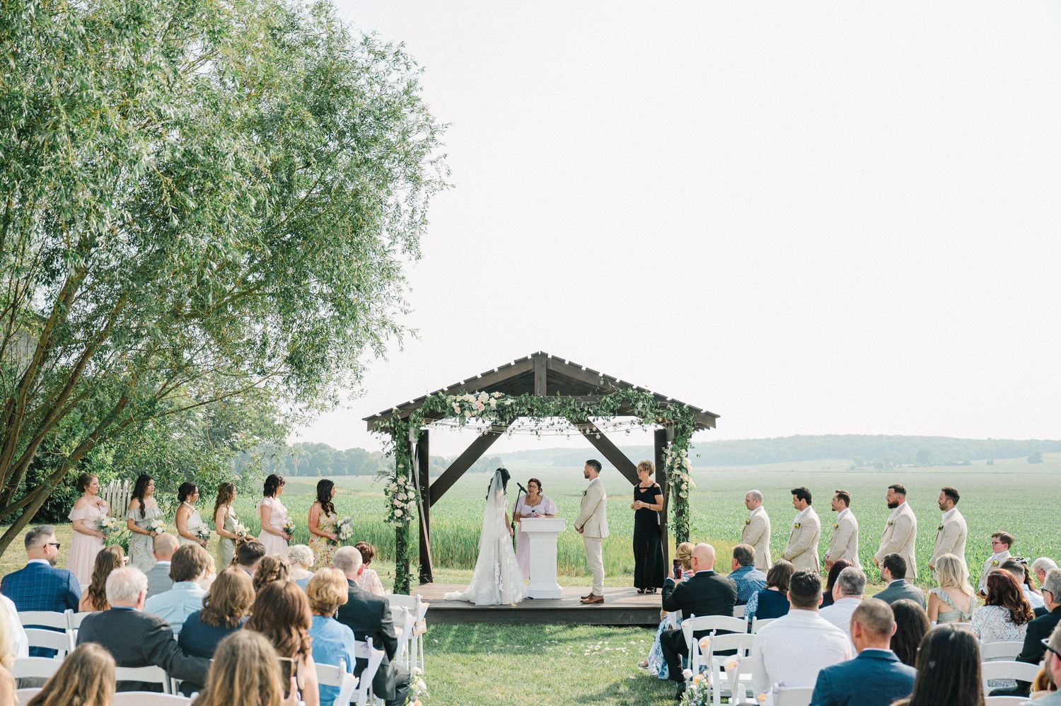 A scenic outdoor wedding ceremony under a rustic wooden arch with rolling hills in the background and guests seated on chairs.