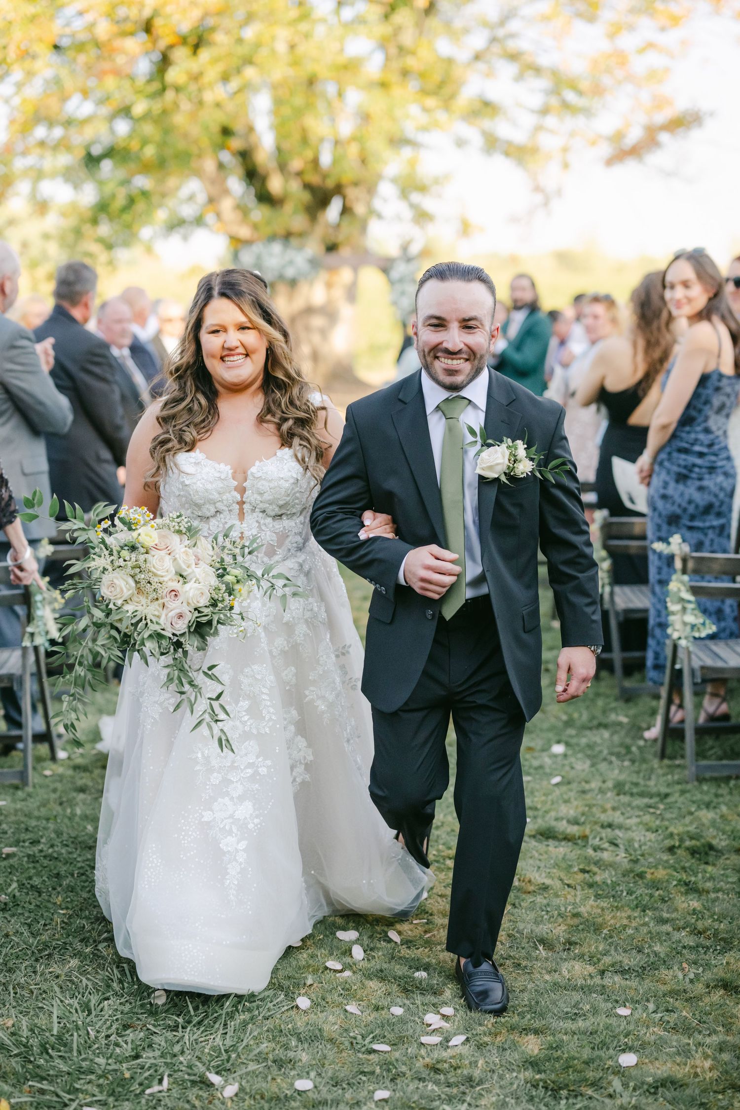 Smiling bride and groom walk down outdoor aisle after wedding ceremony, bride holding white floral bouquet. - Youngblood Vineyard Wedding