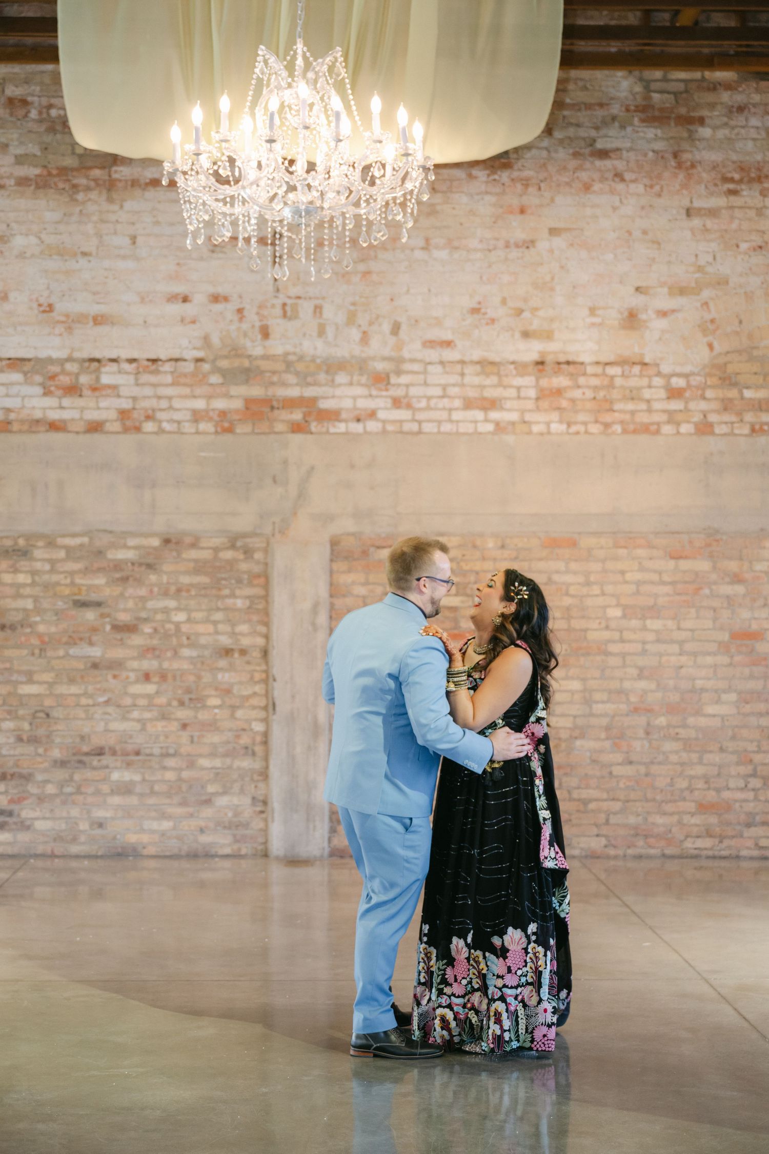 Two people share an intimate moment under elegant crystal chandeliers in a rustic brick-walled venue. - The Brix on the Fox