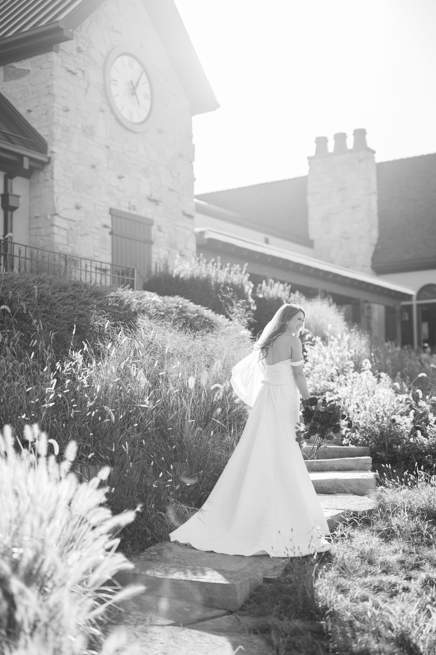 Dreamy black and white photo of a bride standing in a sunlit garden with stone architecture.