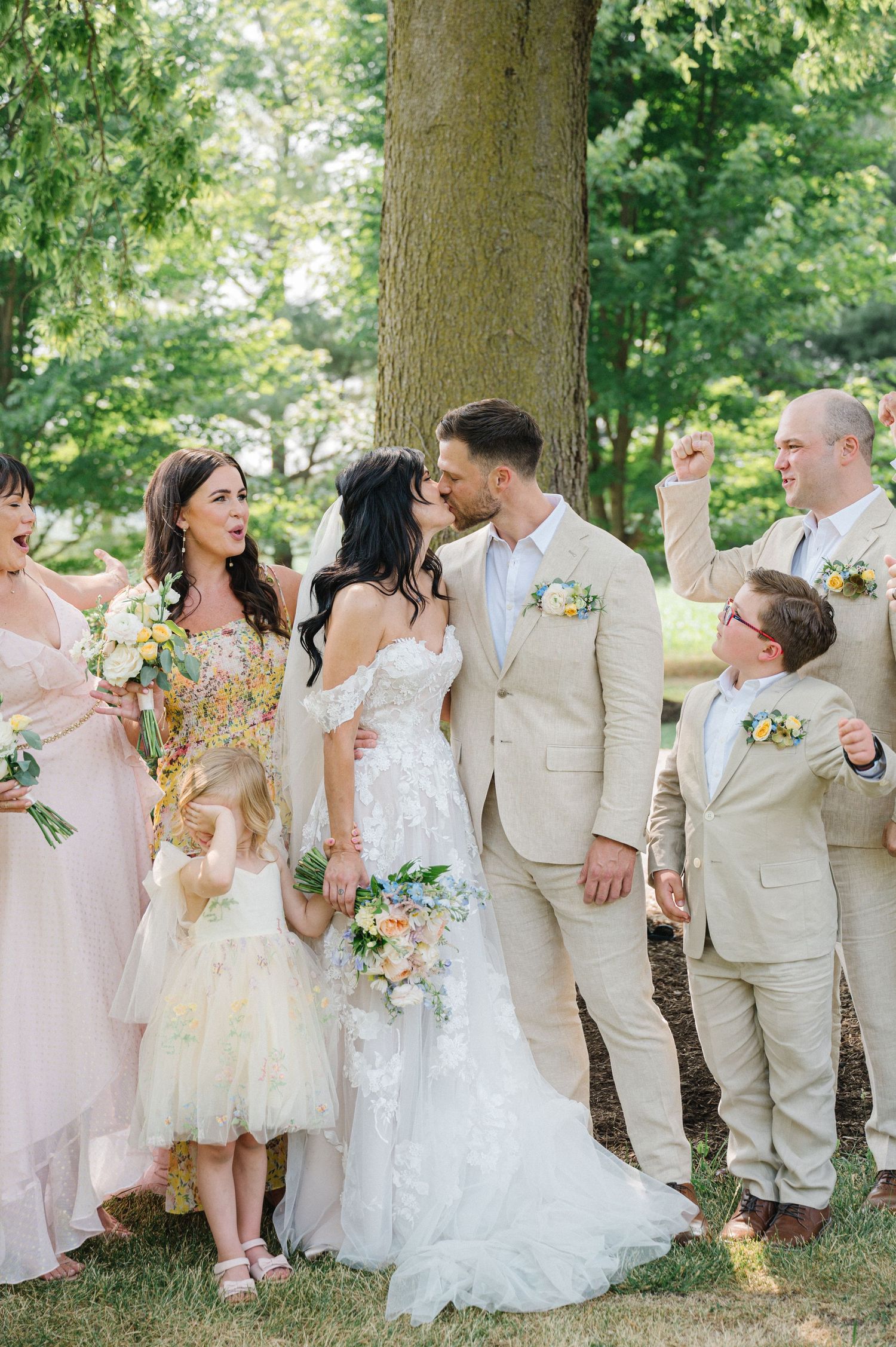 A cheerful wedding party in light colored attire poses together outdoors under a large tree during a summer celebration.