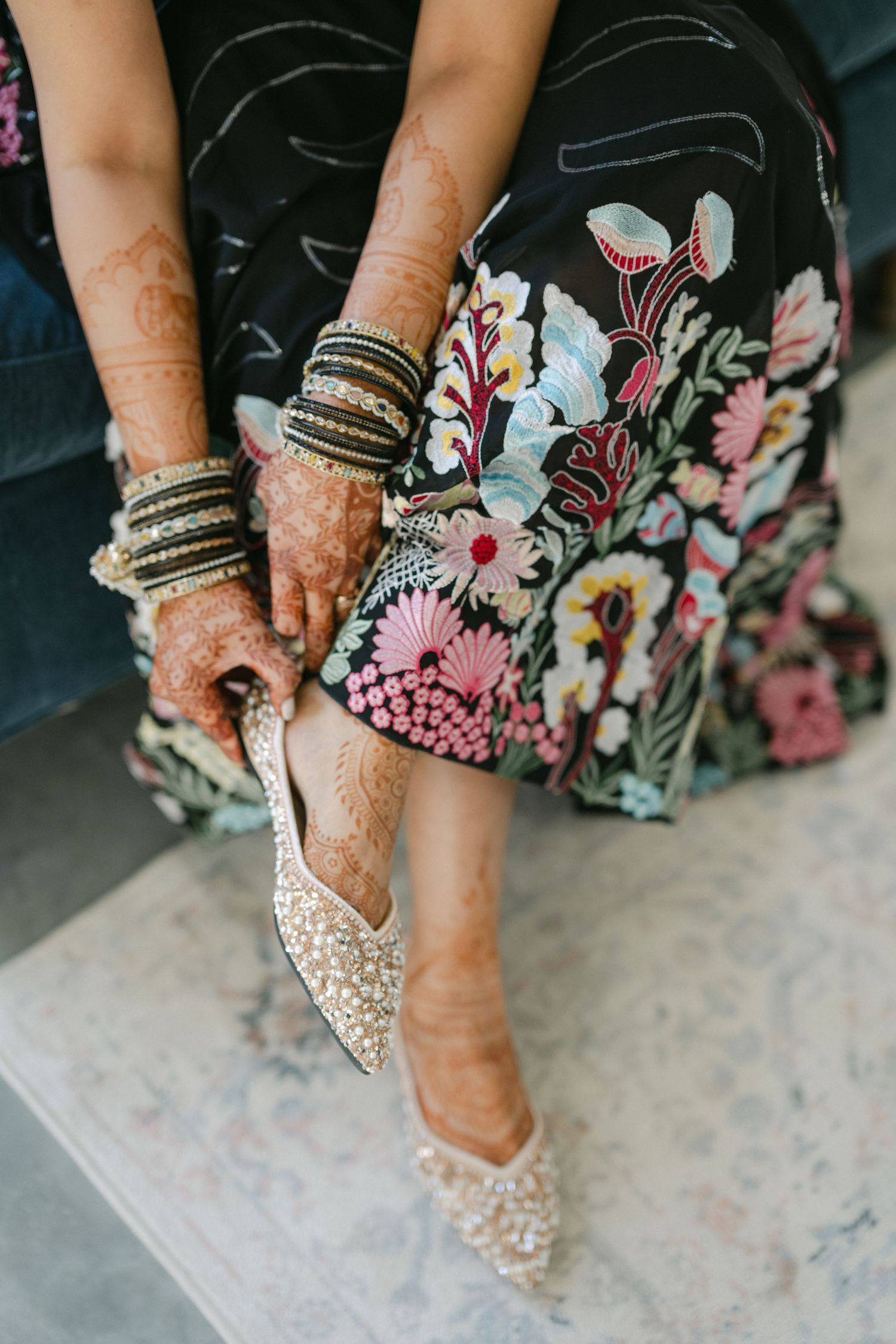 Close-up view of sparkly bridal shoes with henna-adorned feet against floral dress hem on light floor. - The Brix on the Fox