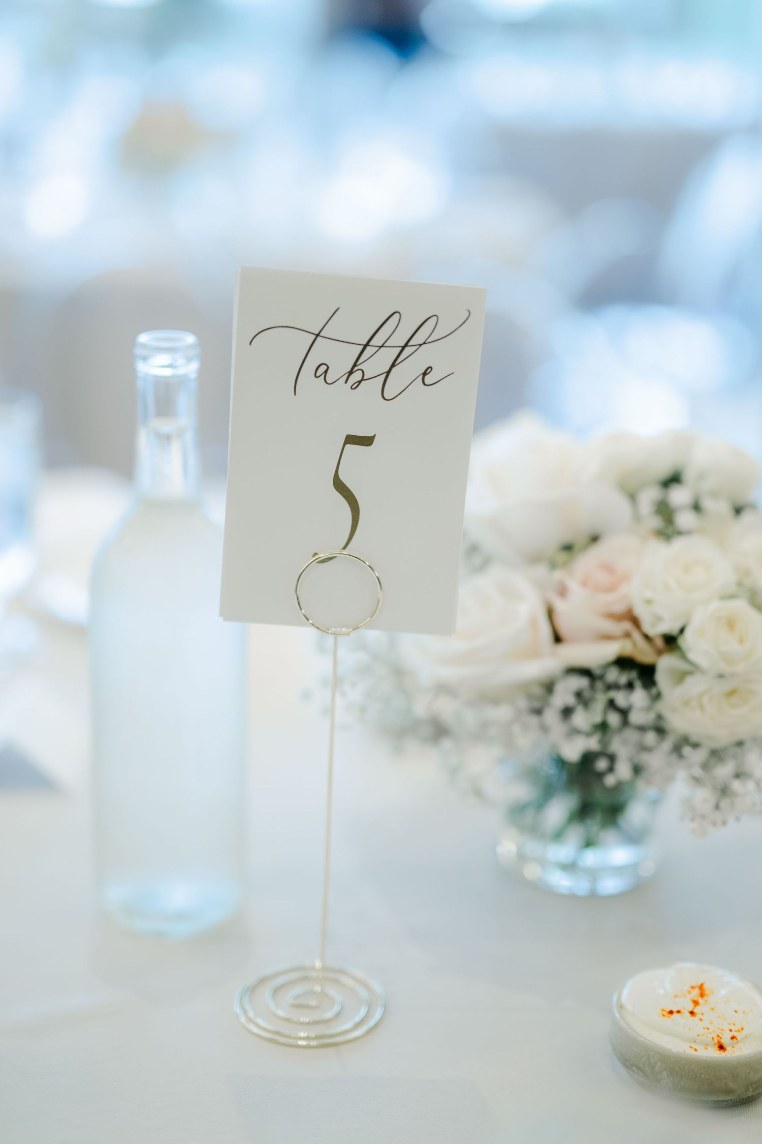 Elegant wedding table number 5 displayed on gold stand with soft white roses and greenery centerpiece in background.  - LM Studio Chicago