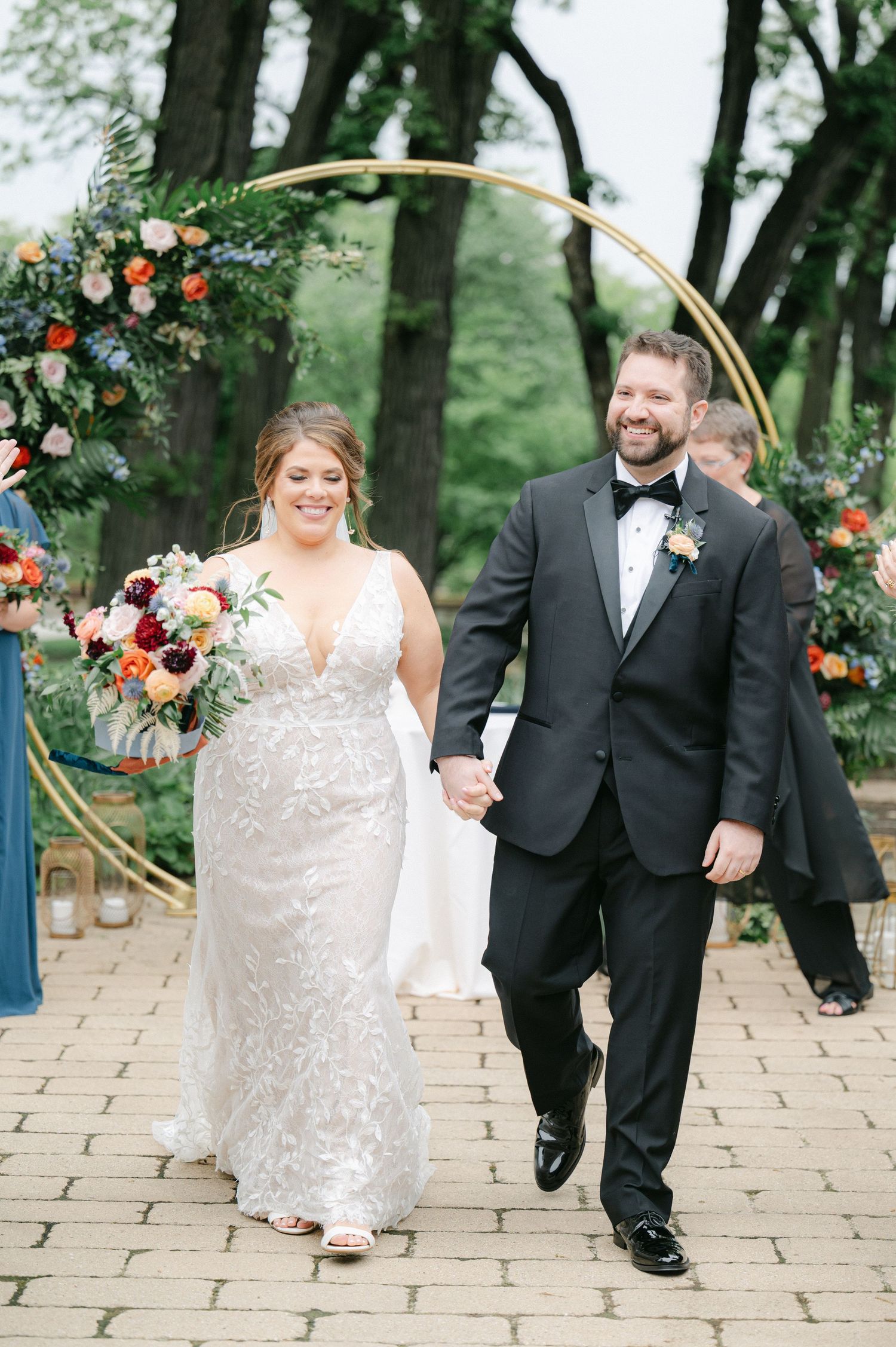 Wedding party walks along stone pathway under trees with floral arch decorations and greenery at outdoor venue. - Hyatt Lodge Oak Brook