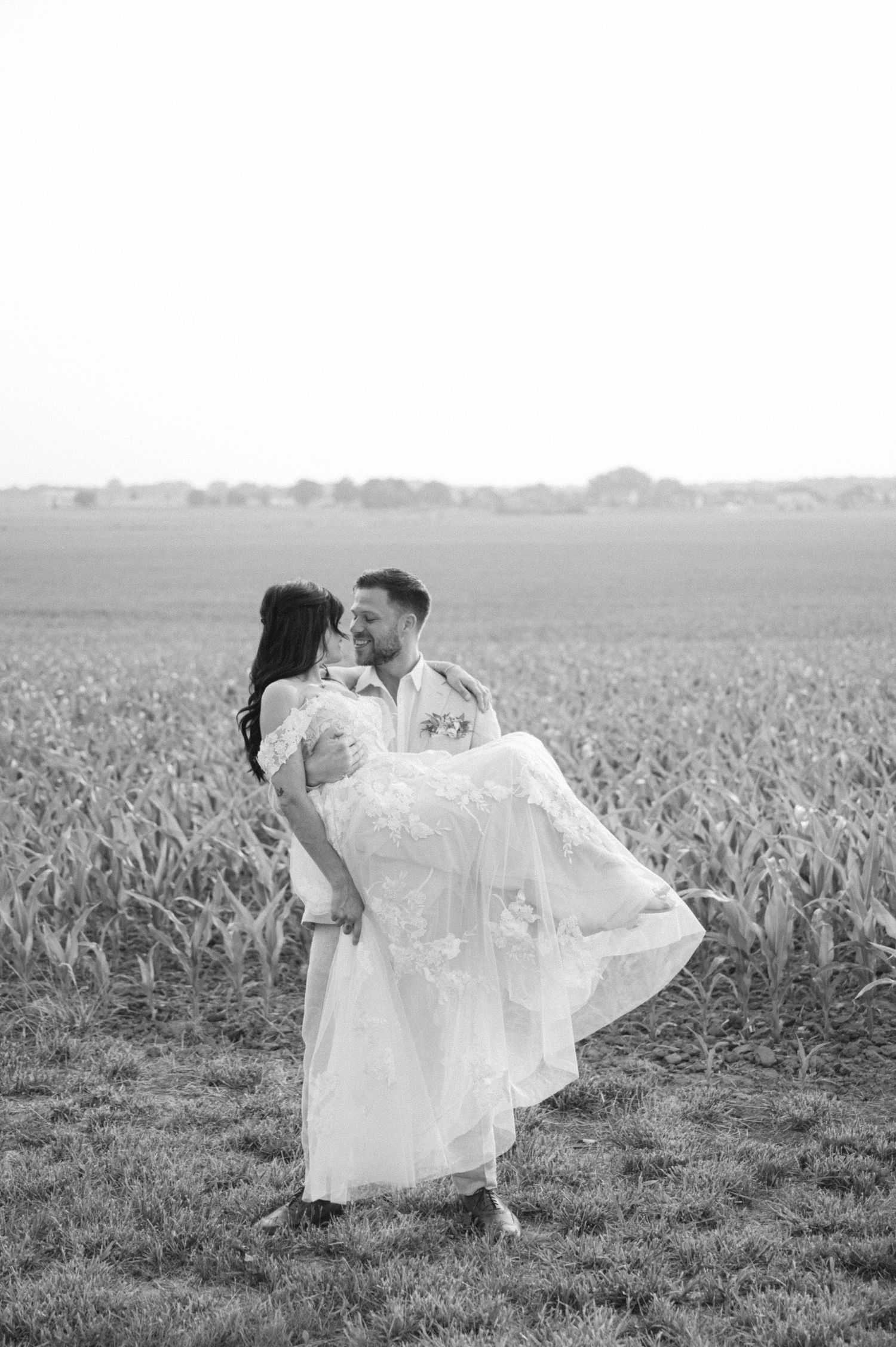 A couple in wedding attire embrace in a scenic field with flowing grass and a misty horizon.