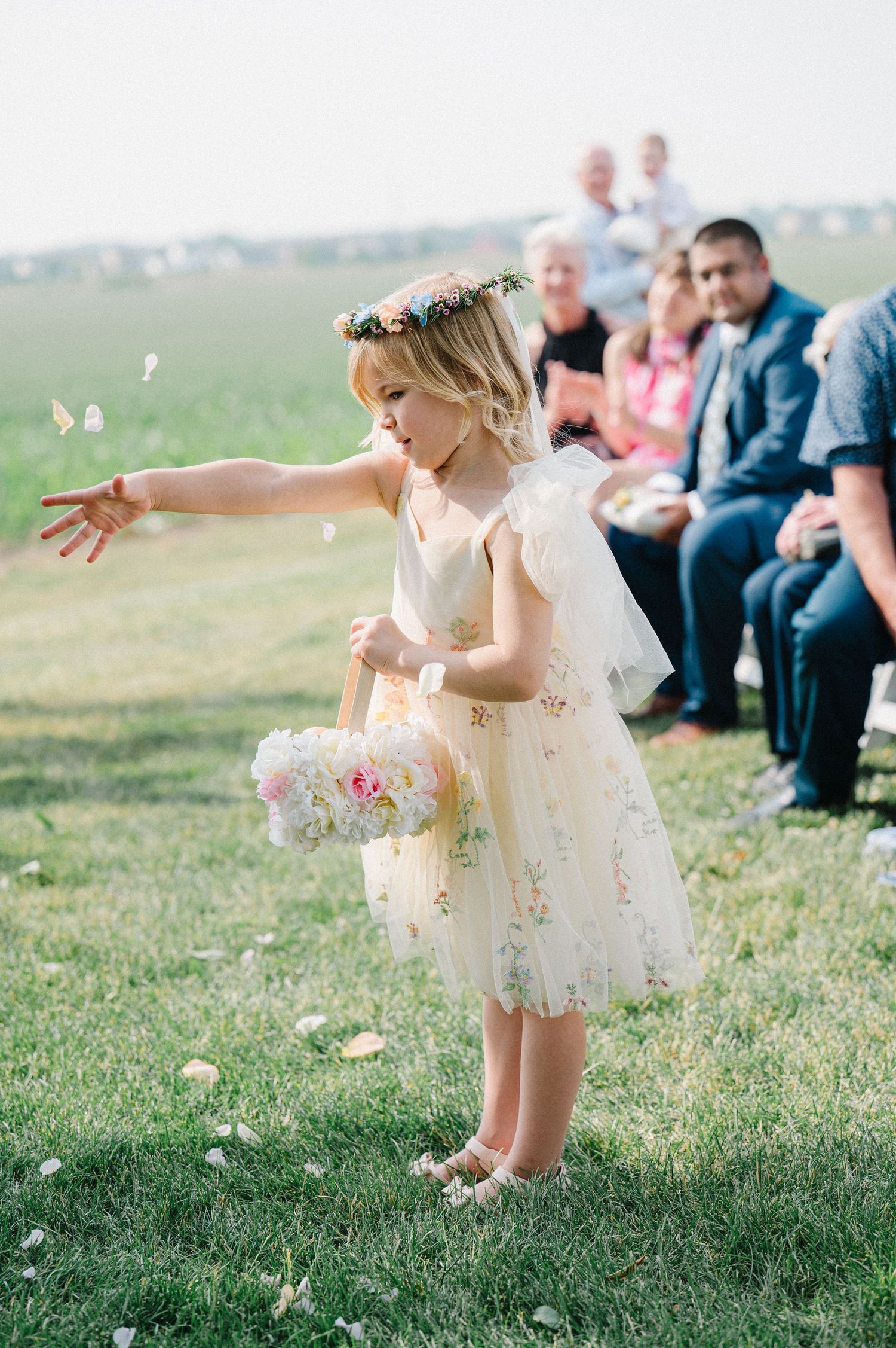 A flower girl in a white lace dress with gold accents tosses petals during an outdoor wedding ceremony on a grassy hill.