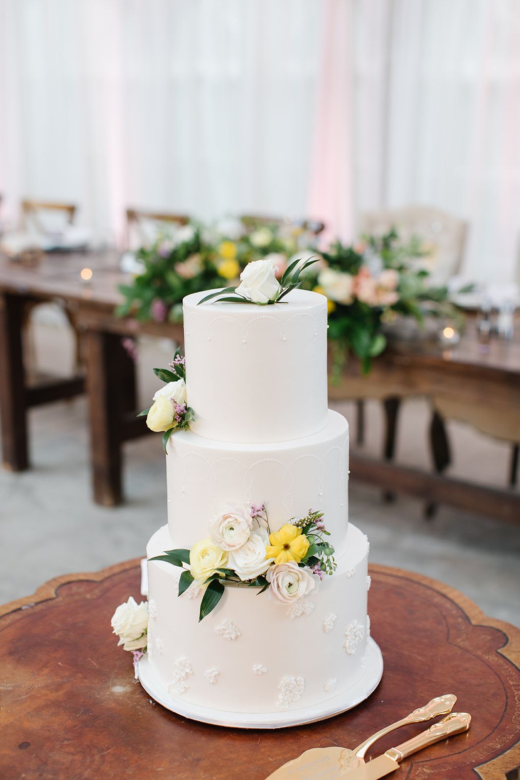 Three-tier white wedding cake with yellow and white floral accents on a wood stand at an elegant reception. - Heritage Prairie Farm Wedding