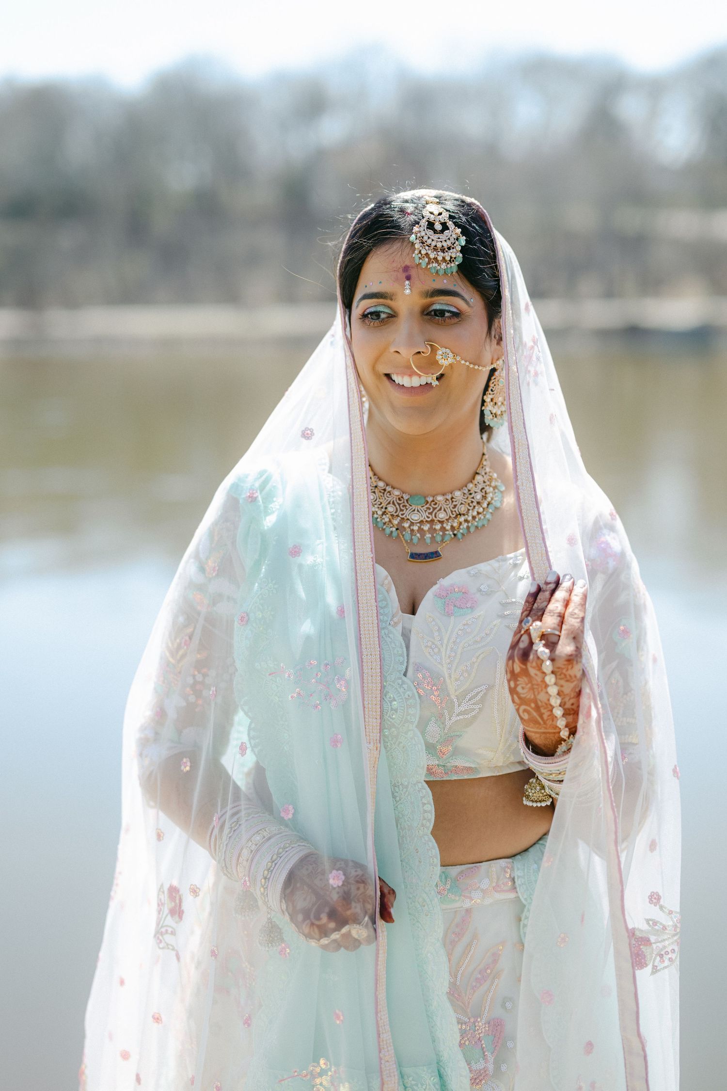 Traditional Indian bride wearing a mint green lehenga with intricate jewelry and veil poses by a lake. - The Brix on the Fox
