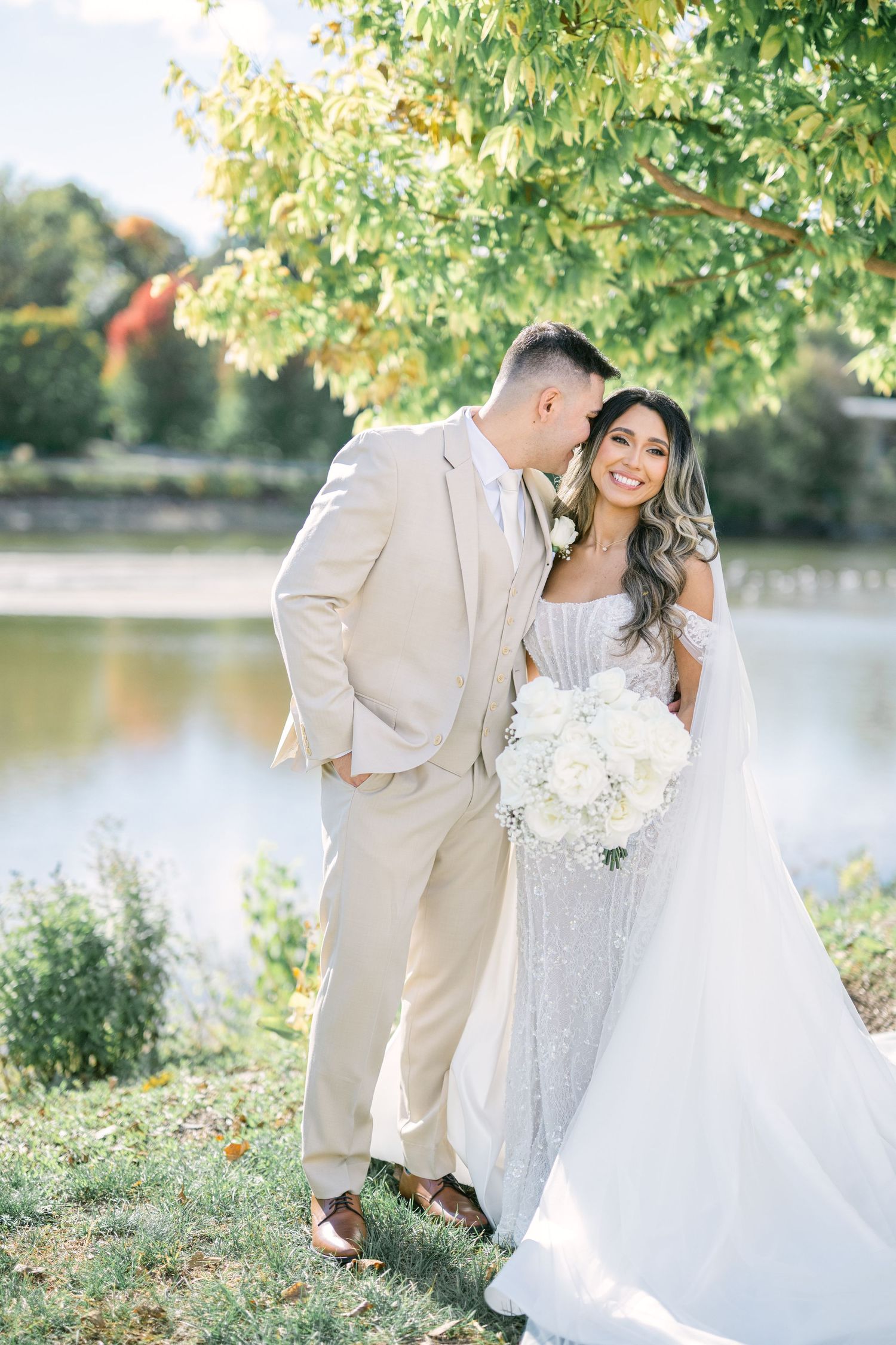 Bride and groom pose by a lakeside under green trees, bride holding white bouquet in elegant gown. - The Brix on the Fox Wedding