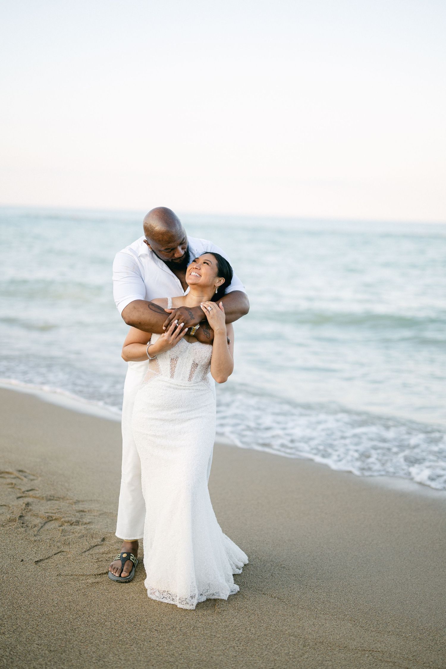 A tender beach wedding moment captured as a couple embraces on the sandy shore beside the ocean.