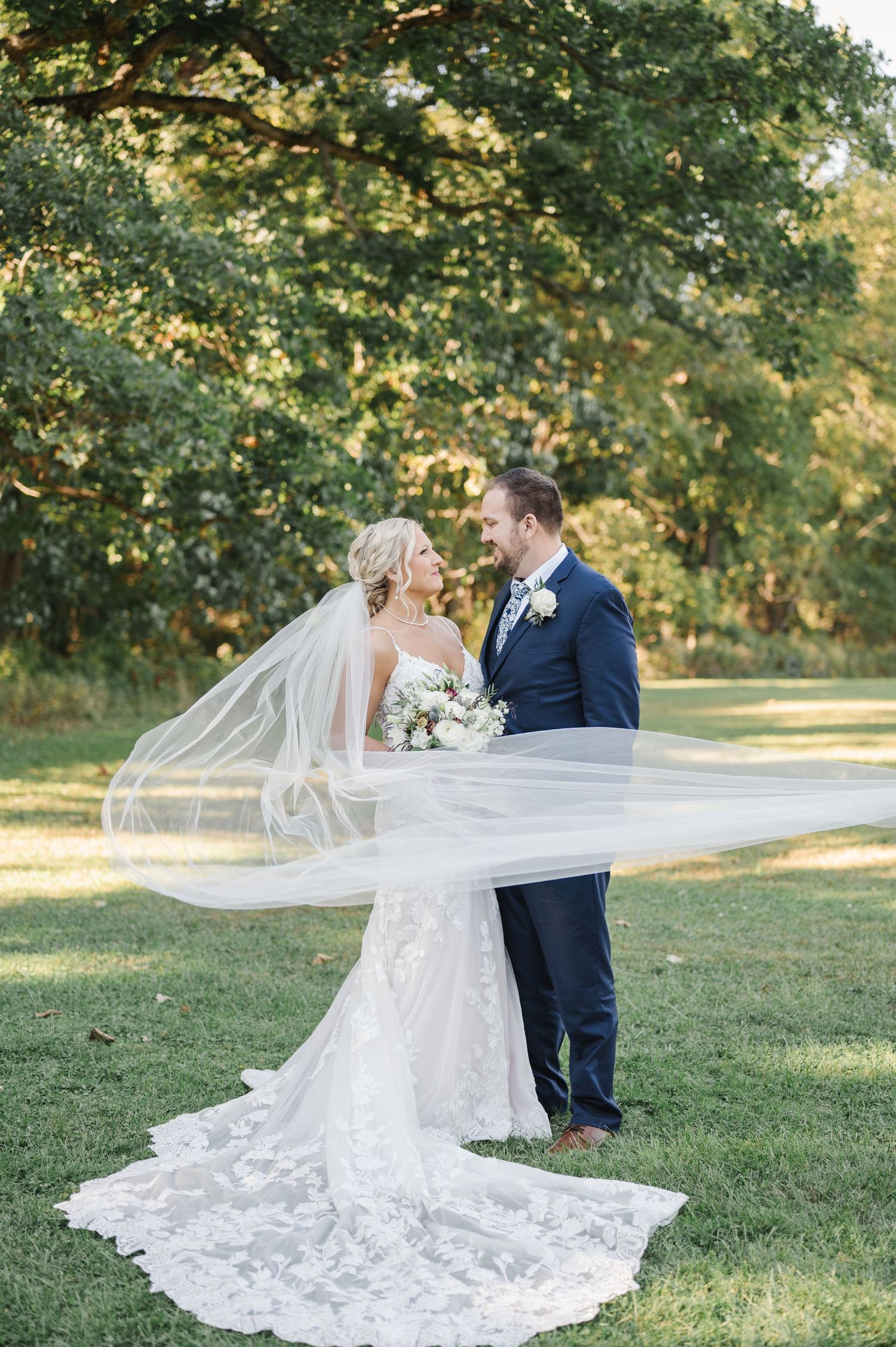 A romantic wedding couple stands together outdoors with a flowing white veil and lace train against a green garden backdrop.