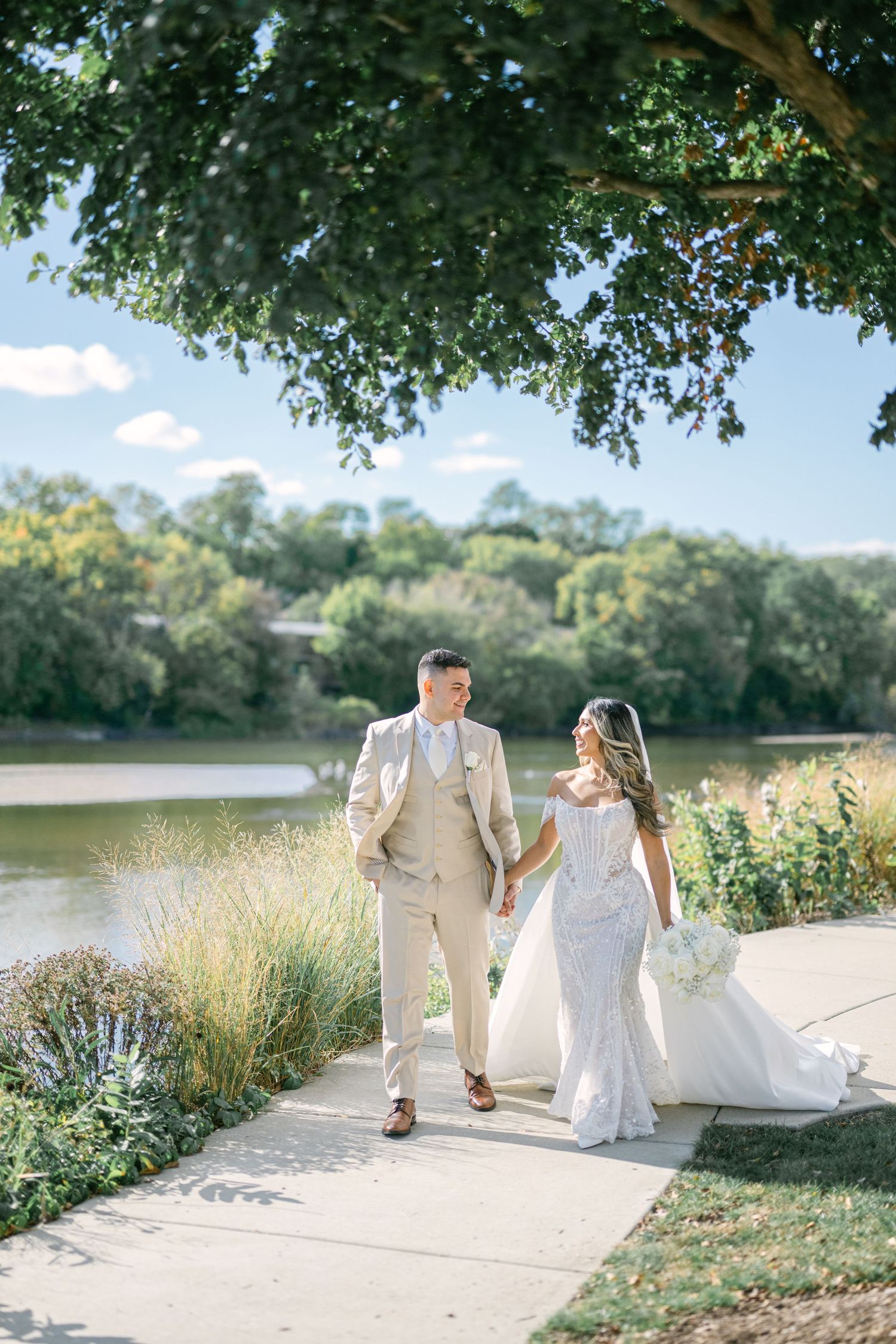 A bride and groom walk hand in hand along a sunlit garden path beside a serene lake on their wedding day. - The Brix on the Fox Wedding