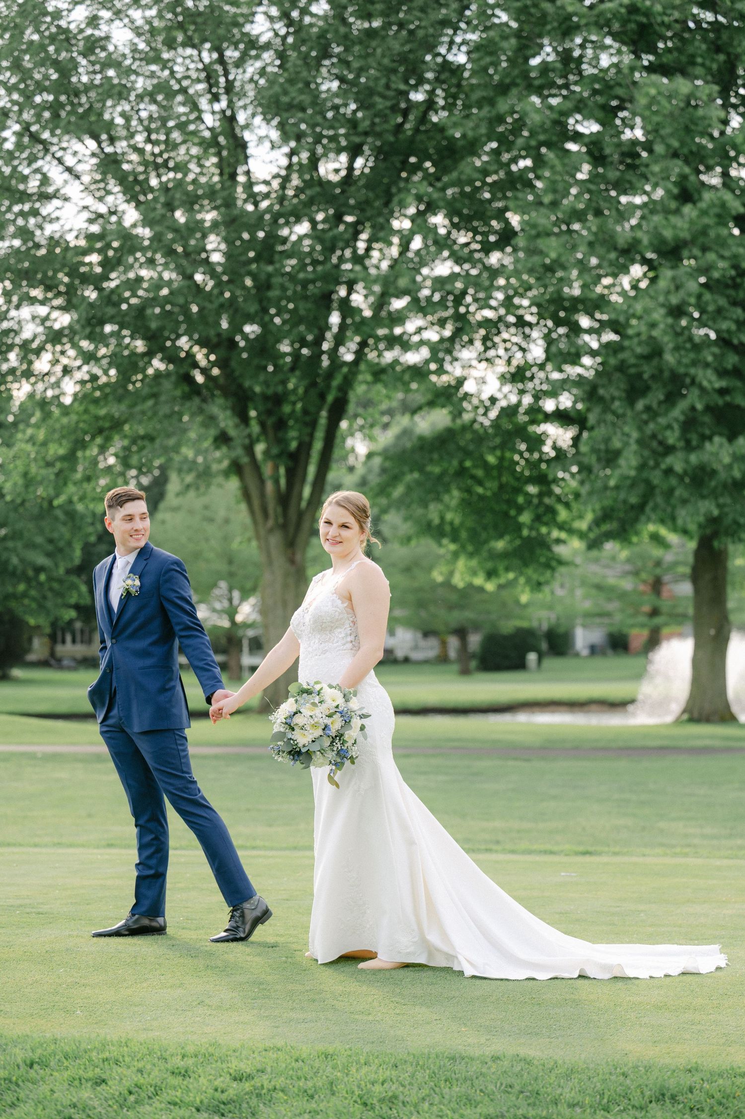 A couple in wedding attire walks hand in hand across a lush green lawn surrounded by tall trees. - Cress Creek Country Club Naperville
