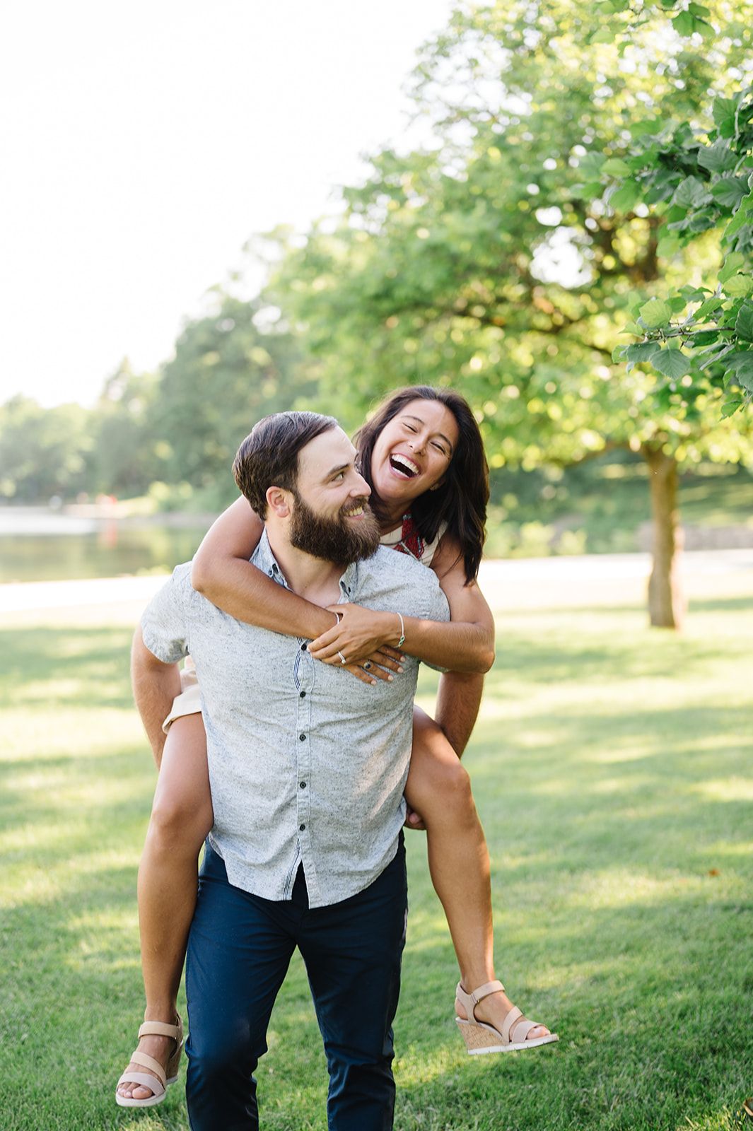 A laughing woman gets a piggyback ride from a bearded man in a sunny green park setting. - Wheaton Engagement Session