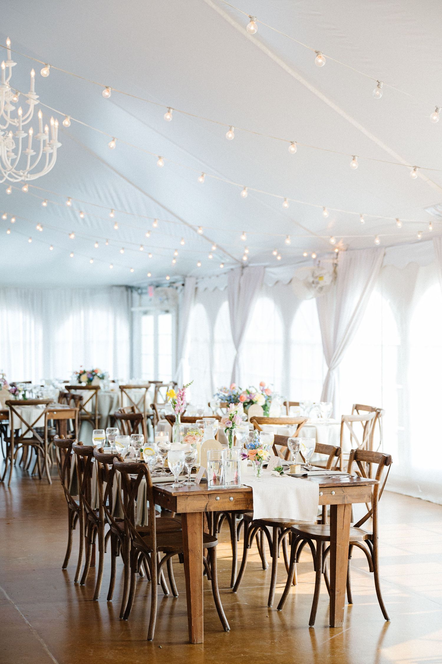 Elegant wedding reception venue with white drapes, twinkling lights, wooden cross-back chairs and farm tables under a chandelier.