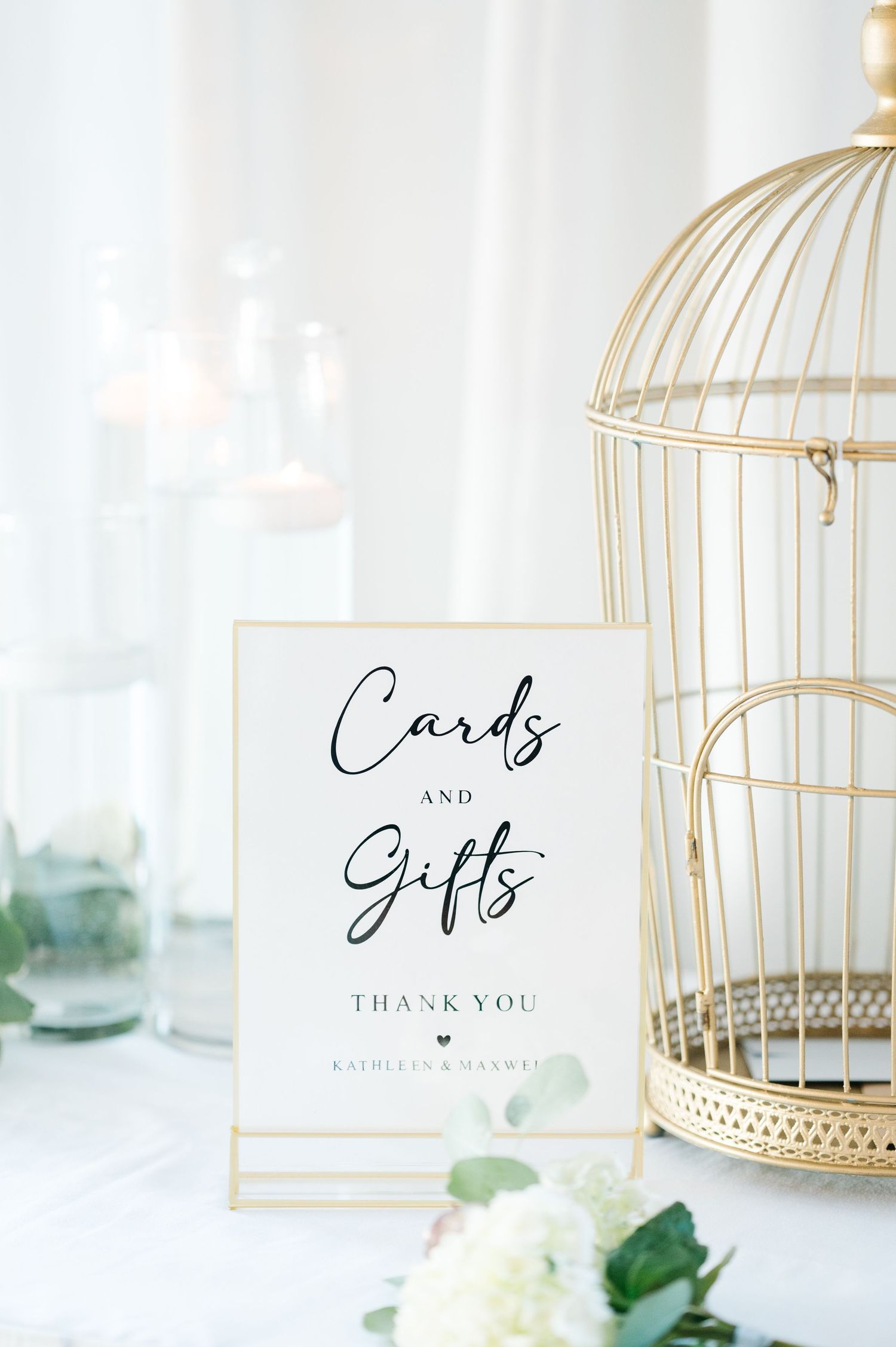 Elegant wedding cards and gifts sign displayed next to a gold birdcage and white roses on a white table.