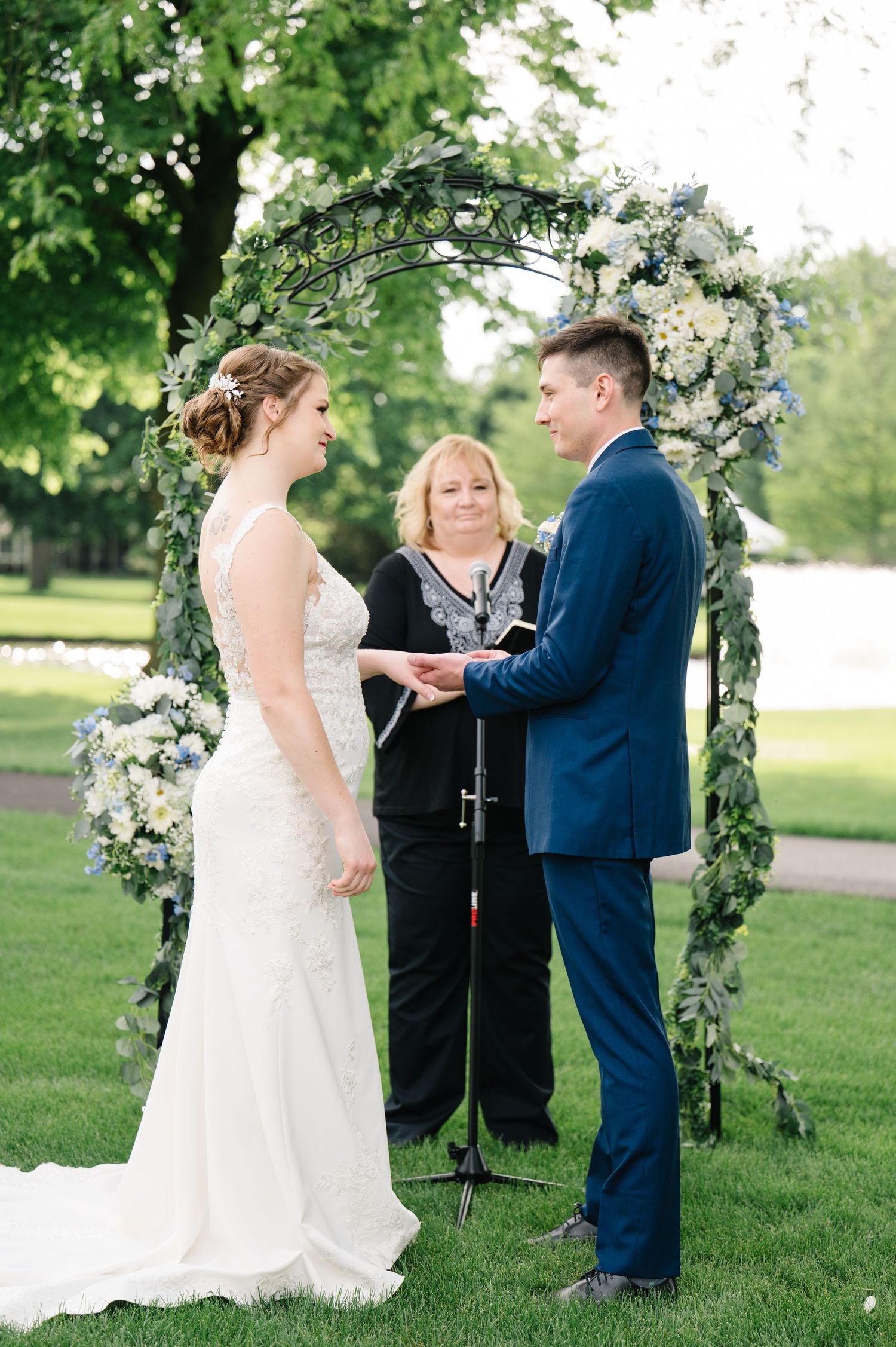 An outdoor wedding ceremony under a floral arch decorated with white flowers and greenery in a lush garden setting.